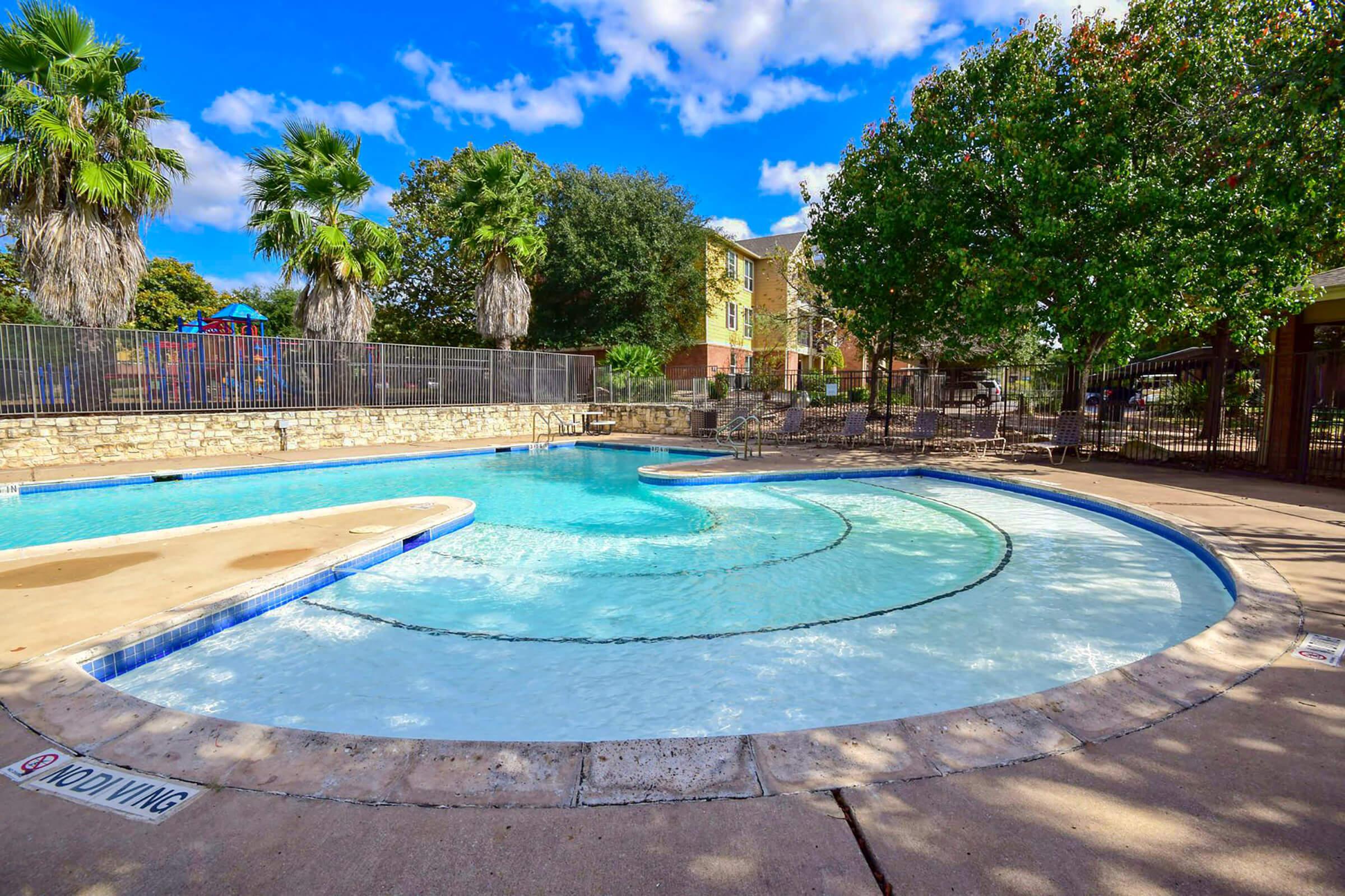 A clear, inviting swimming pool with a curved design, surrounded by palm trees and lush greenery. The pool area features a smooth concrete deck, and nearby, there is a fence for safety. Bright blue skies with scattered clouds add to the serene atmosphere.