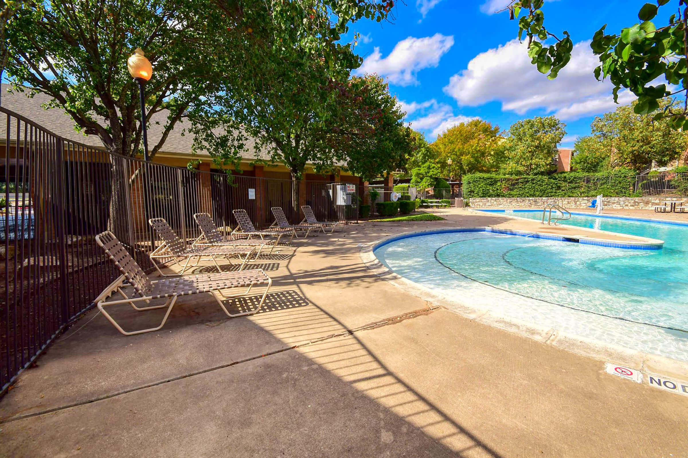 A tranquil outdoor swimming pool area with clear water, surrounded by a black wrought-iron fence. Several lounge chairs are positioned on the concrete deck, with shade from nearby trees. The sky is blue with fluffy white clouds, and the pool features a curved design with steps leading into the water.