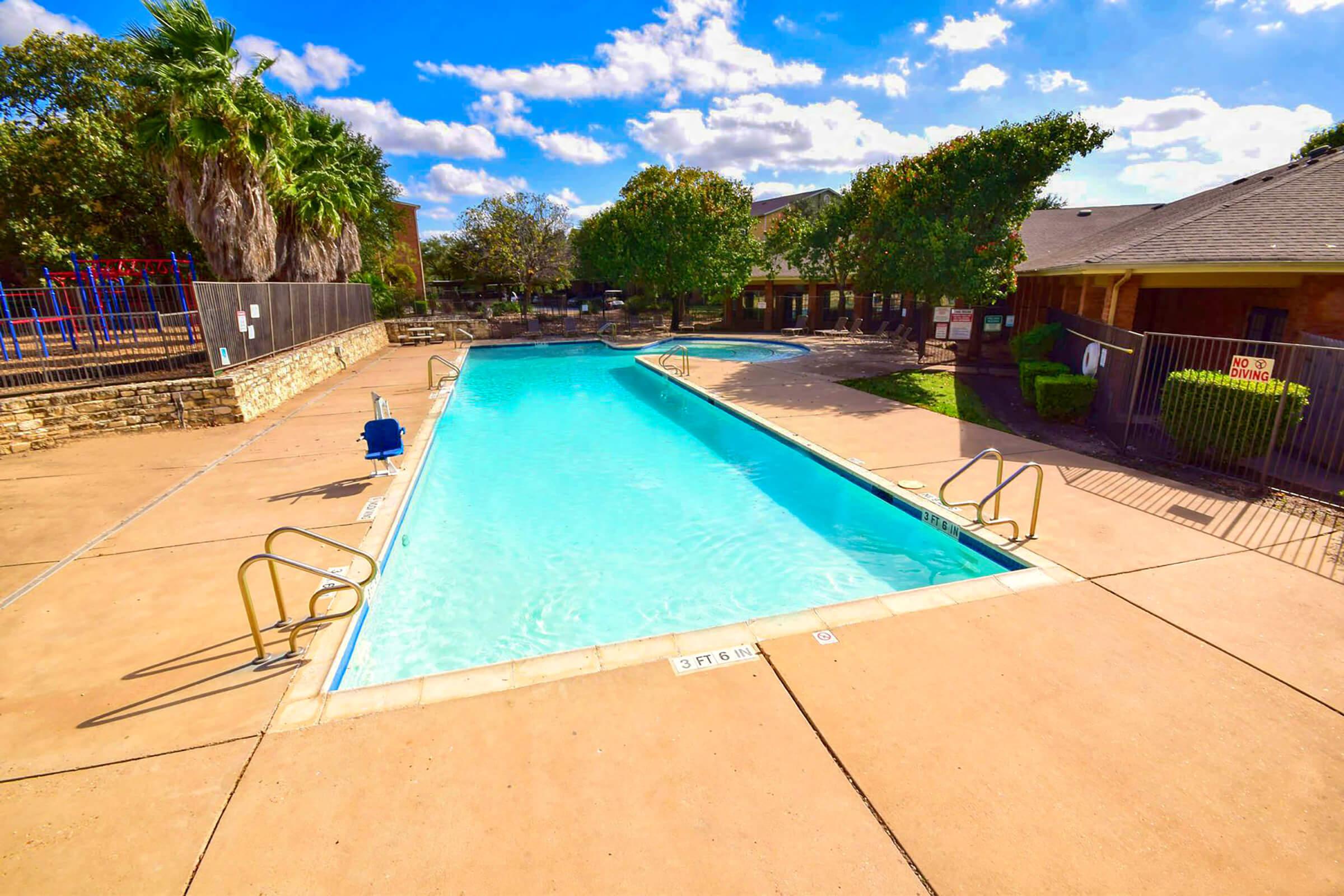A clear swimming pool under a bright blue sky with scattered clouds. On one side, there are lounge chairs and a small area with playground equipment. Surrounding the pool area, there are trees providing shade and a fenced perimeter with a few buildings in the background. The scene is inviting and sunny.