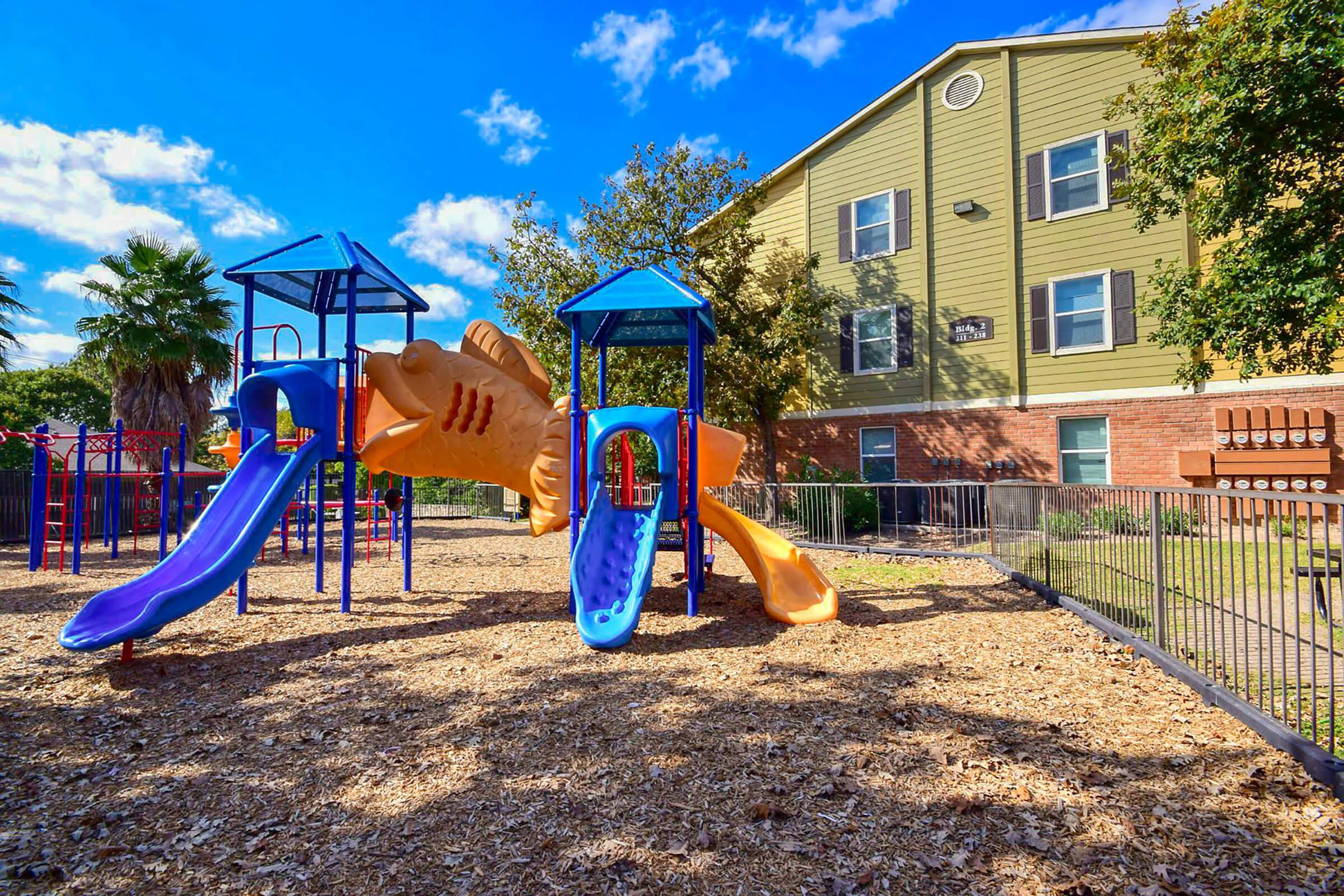 A colorful playground featuring two slides and a fish-shaped climbing structure, surrounded by wood chips. In the background, there is a green multi-family building with windows and greenery, under a clear blue sky with fluffy clouds.