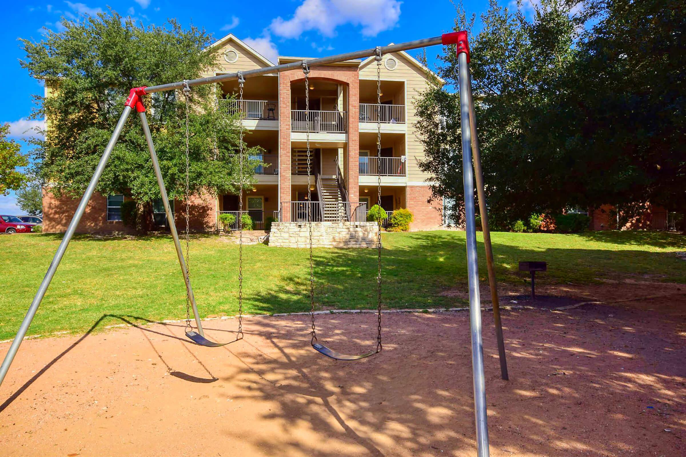 A playground with metal swings in front of a multi-story brick apartment building surrounded by grass and trees. Blue sky with scattered clouds is visible in the background.