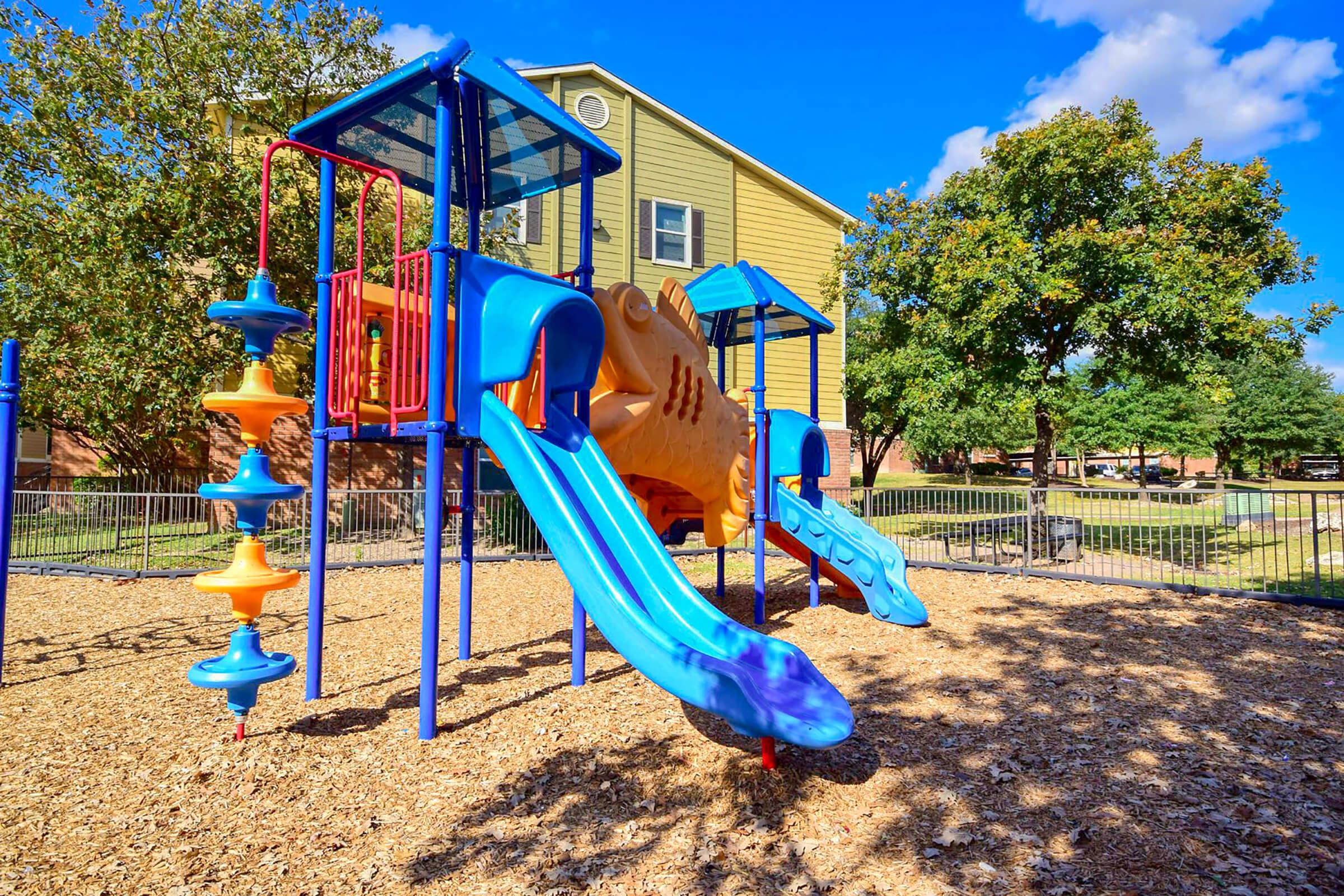 Colorful playground equipment featuring two large slides, jungle gym, and climbing structures, set on a bed of wood chips. In the background, a yellow building and green trees are visible under a blue sky with fluffy clouds. The area is fenced for safety.