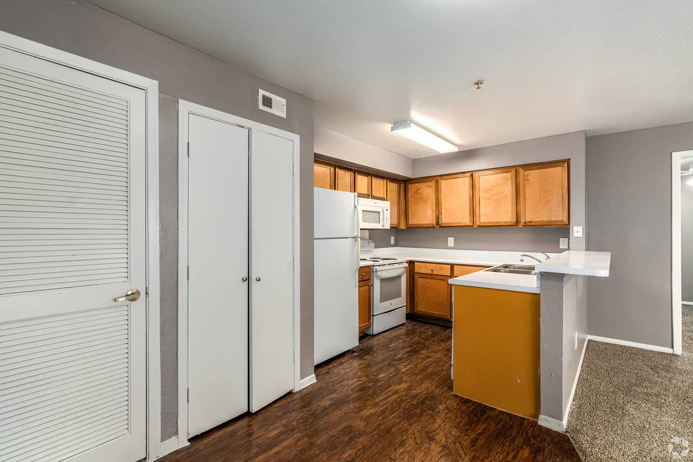 A modern kitchen featuring wooden cabinets, a white refrigerator, and a stove. The space includes a breakfast bar, light gray walls, and laminate flooring. Storage closets are visible next to the entrance, providing an organized and functional layout.