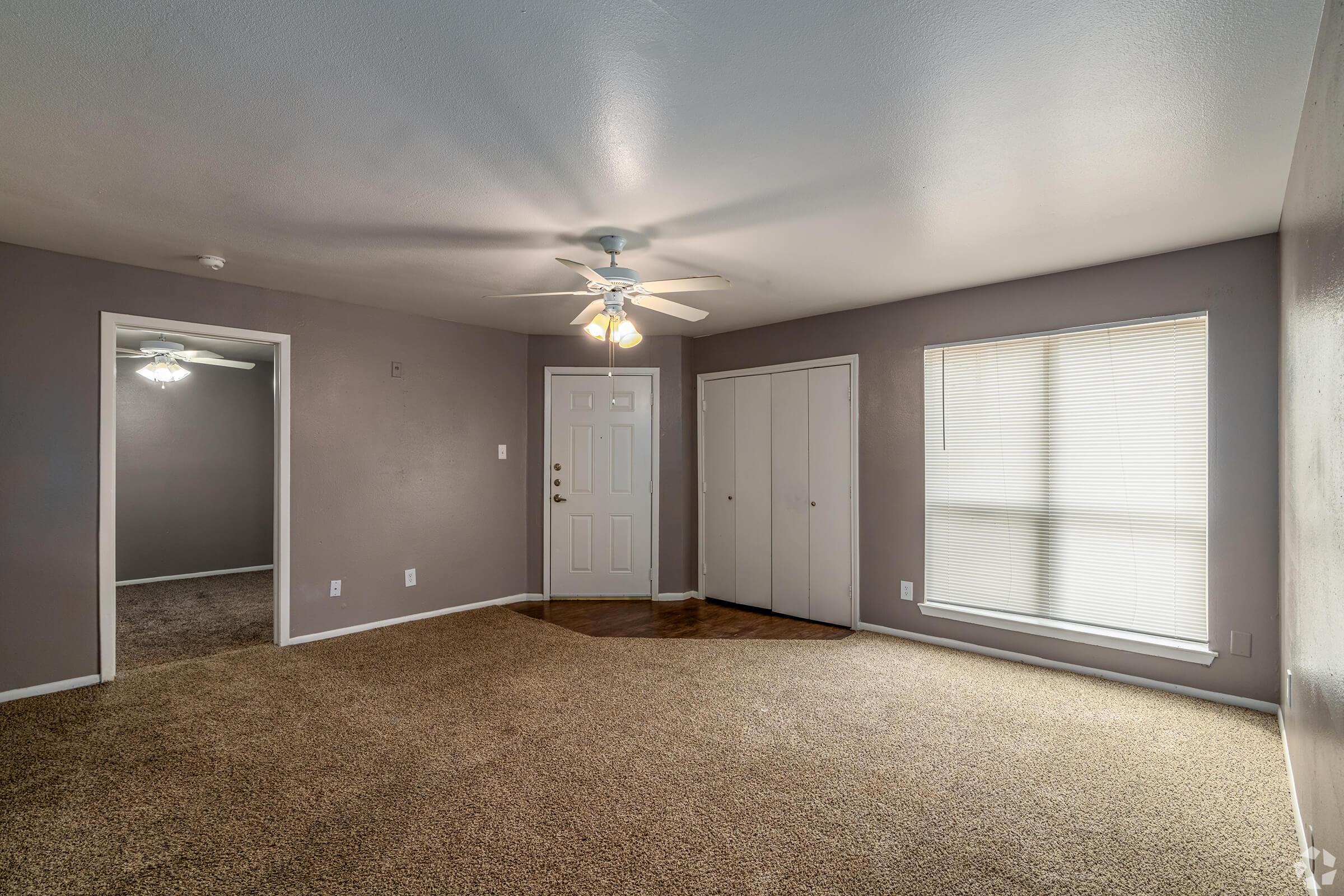 A spacious living room with light gray walls and carpeted flooring. The room features a ceiling fan, a front door, a closet with sliding doors, and a large window with blinds. An adjoining room is visible through an open doorway on the left. Overall, the space is well-lit and inviting.