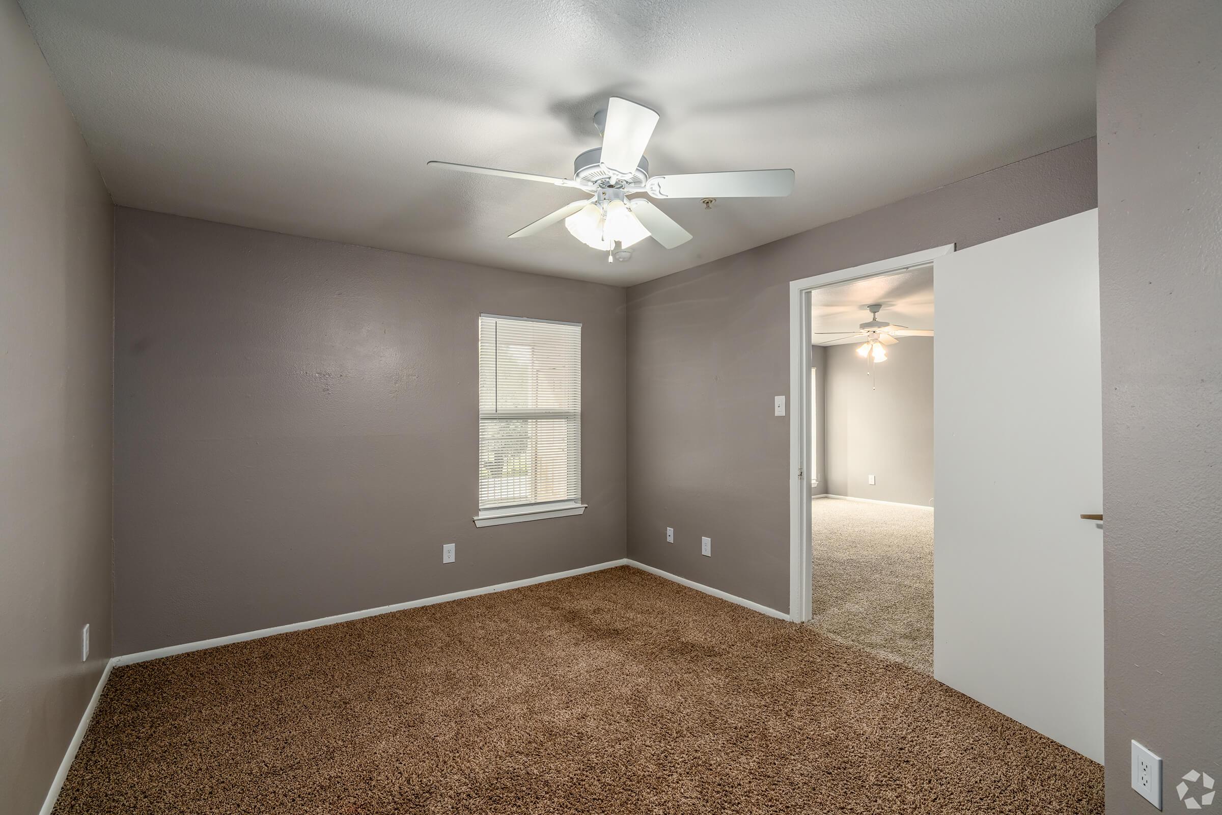 A spacious, neutral-colored room with a light brown carpet and a ceiling fan. The walls are painted a soft gray, and there is a window letting in natural light. A door on the right leads to another room. The overall ambiance is bright and inviting, suitable for various uses.