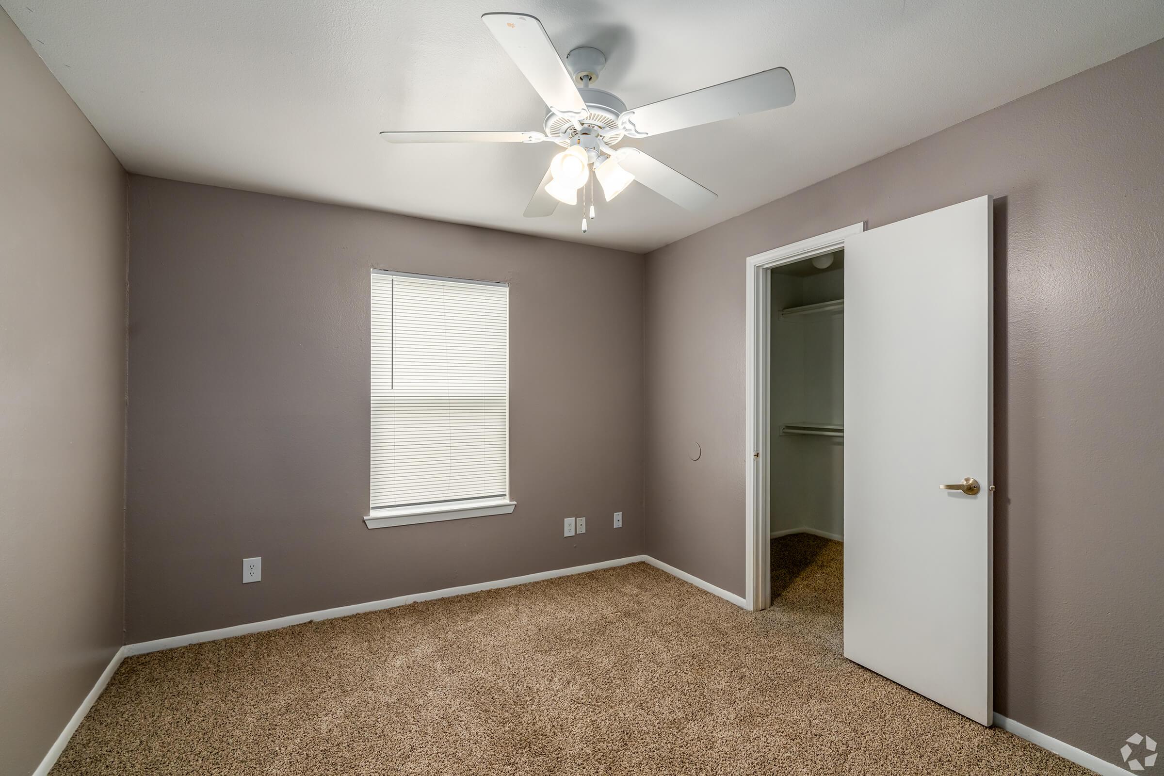 A small, empty room with light brown carpet and gray walls. A ceiling fan with four light bulbs is mounted in the center. There is a window with white blinds on the left wall and a white door leading to a closet on the right. The room has a simple, unadorned appearance, suggesting it is ready for customization.