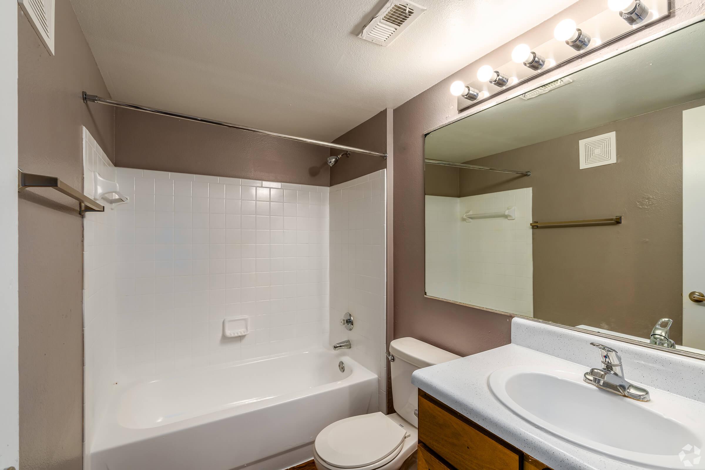 A clean and modern bathroom featuring a white tiled bathtub with a shower, a white countertop with a sink, a large mirror above the sink, and light fixtures. The walls are painted a warm neutral color, and there is a towel rack and a small ventilation grill in the ceiling.