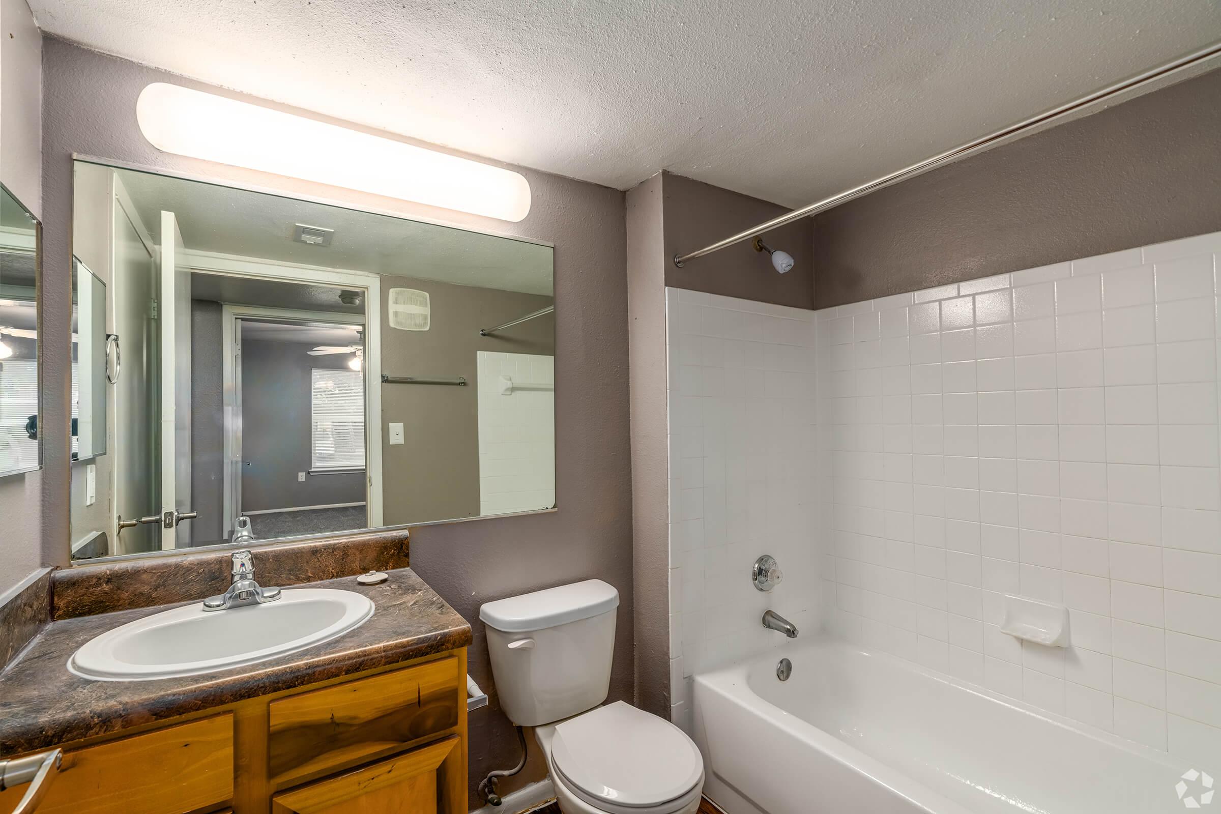 A clean and simple bathroom featuring a white tub and shower, a wooden vanity with a sink, and a mirror above it. The walls are painted in a soft gray color. There is a toilet next to the vanity, and a sliding door leads to another room. The overall design is functional and well-lit.