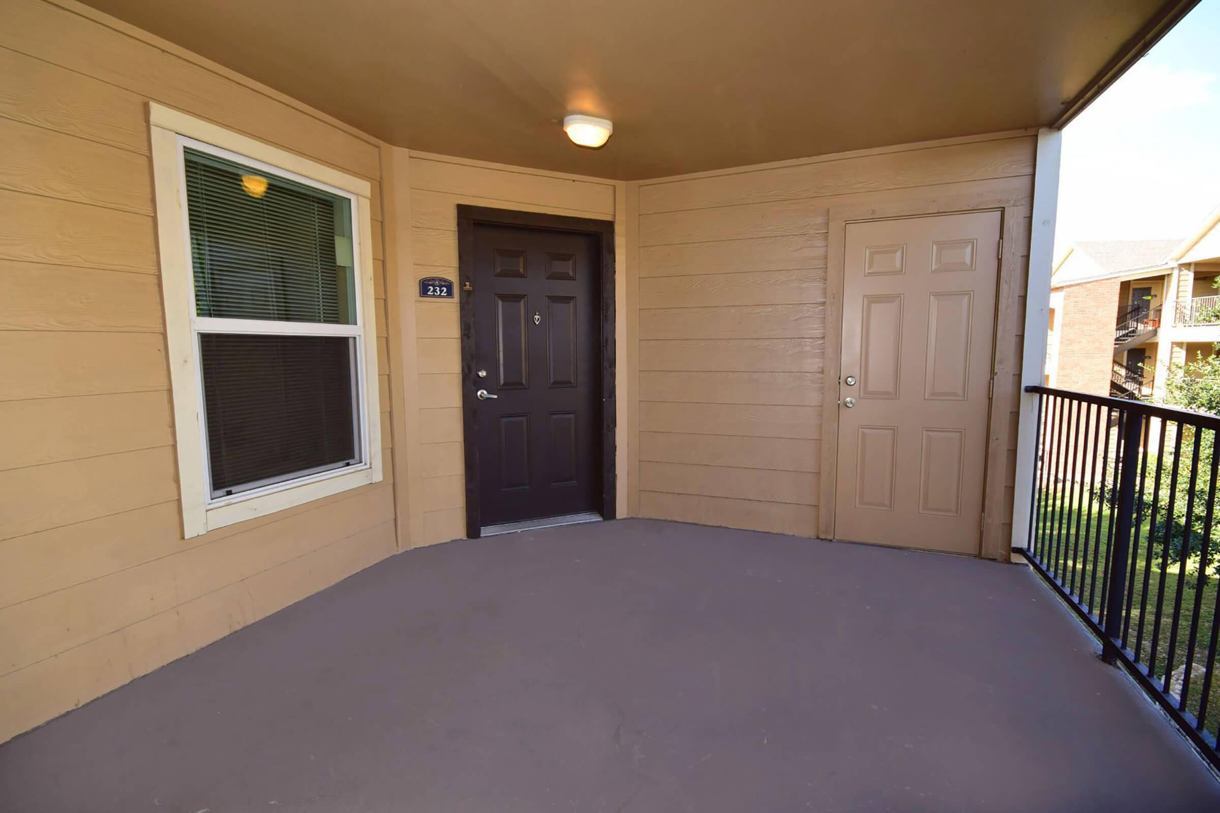 A well-lit entryway to an apartment with a dark brown door and a light beige exterior wall. There is a window to the left and a neighboring door on the right, both contributing to a welcoming atmosphere. The floor is smooth and unadorned, providing a spacious feel to the entrance.