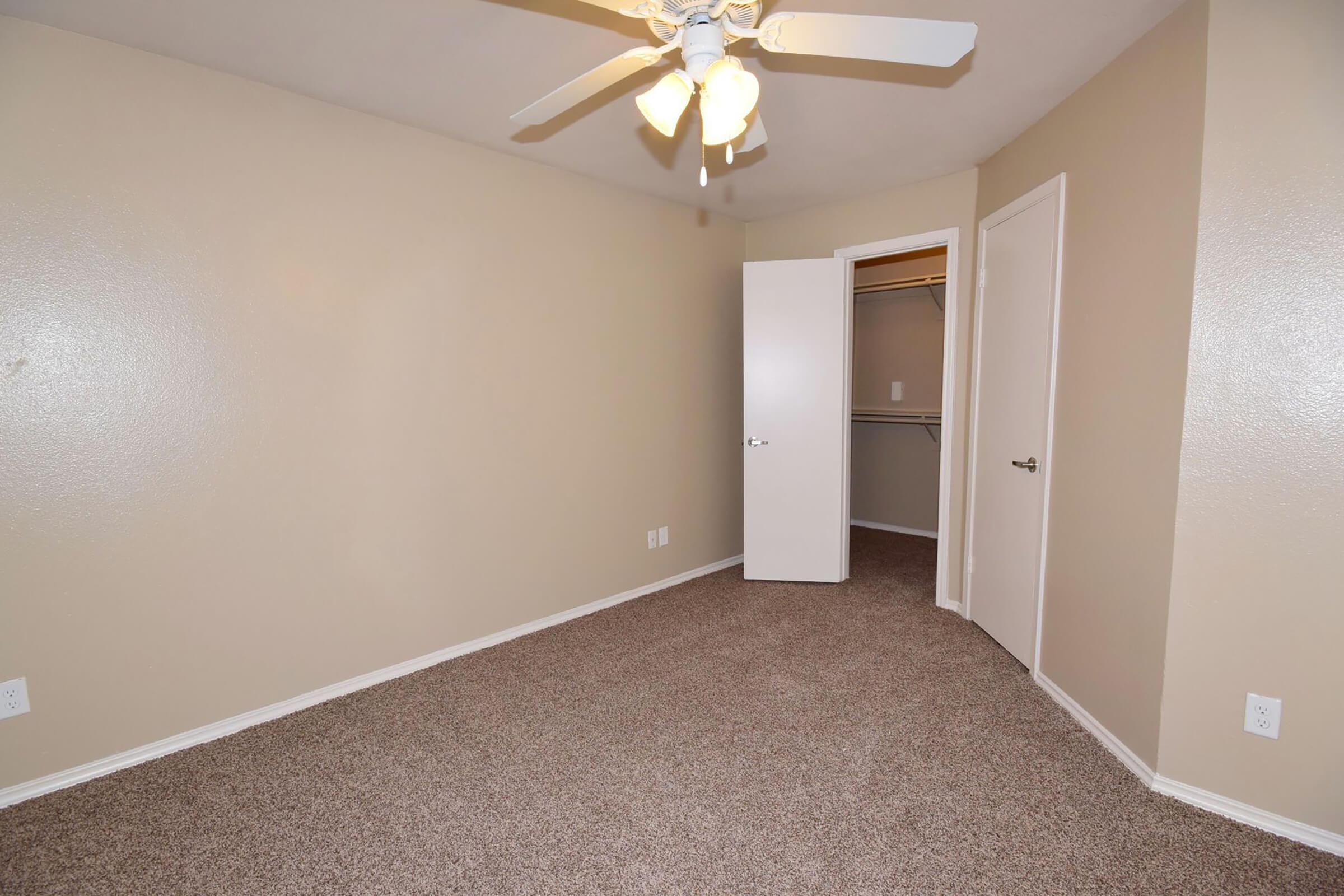 A vacant room with light beige walls and brown carpet. There is a ceiling fan with four lights in the center. One door is slightly ajar, revealing a closet with a shelf. The room has no furniture, giving it a spacious, empty appearance.