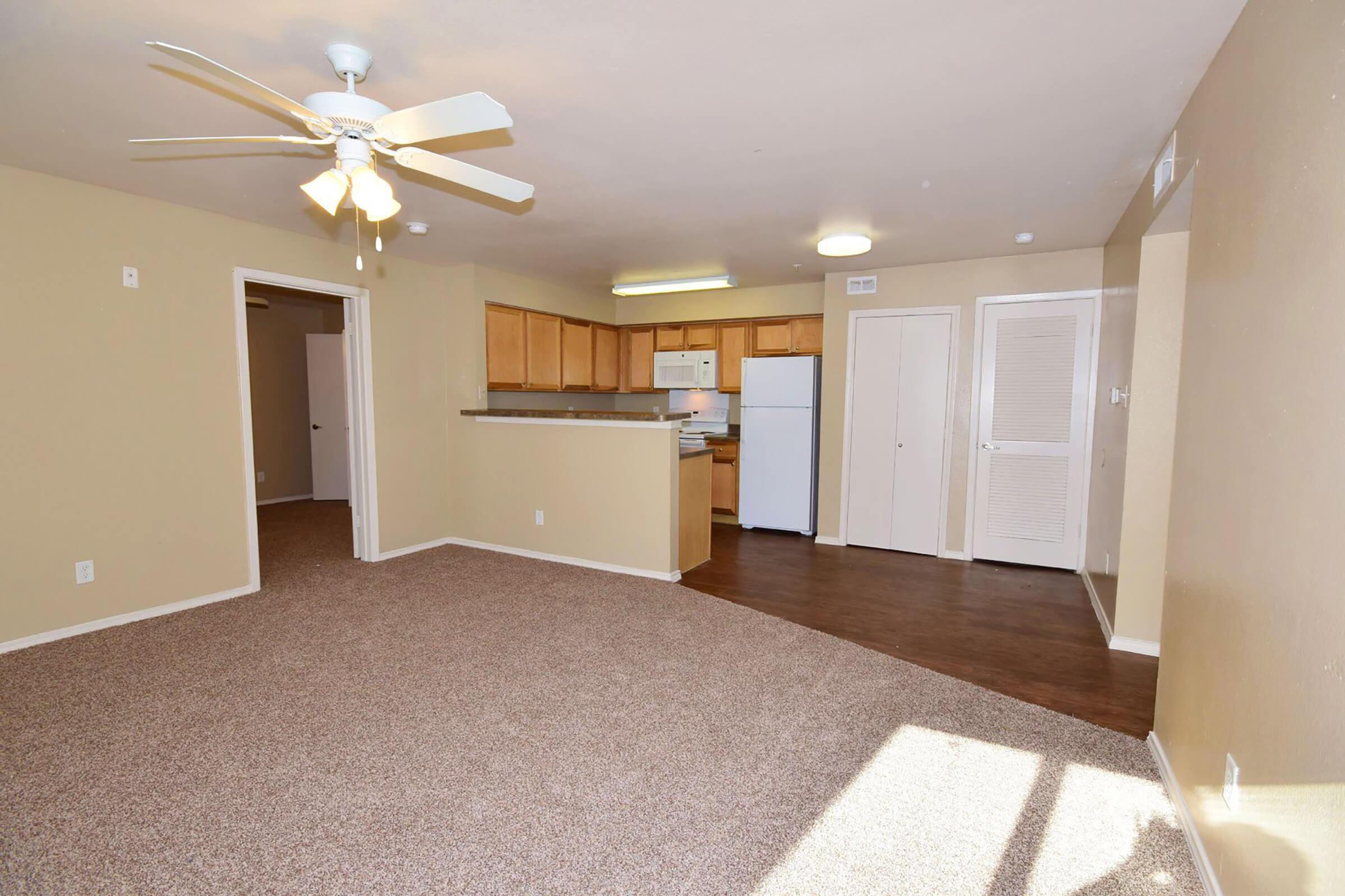Spacious living area with beige walls and carpet, featuring a ceiling fan. A kitchen is visible in the background with wooden cabinets and white appliances. There is a doorway leading to another room and a closet with sliding doors, creating an inviting and bright atmosphere. Natural light streams in from a window.