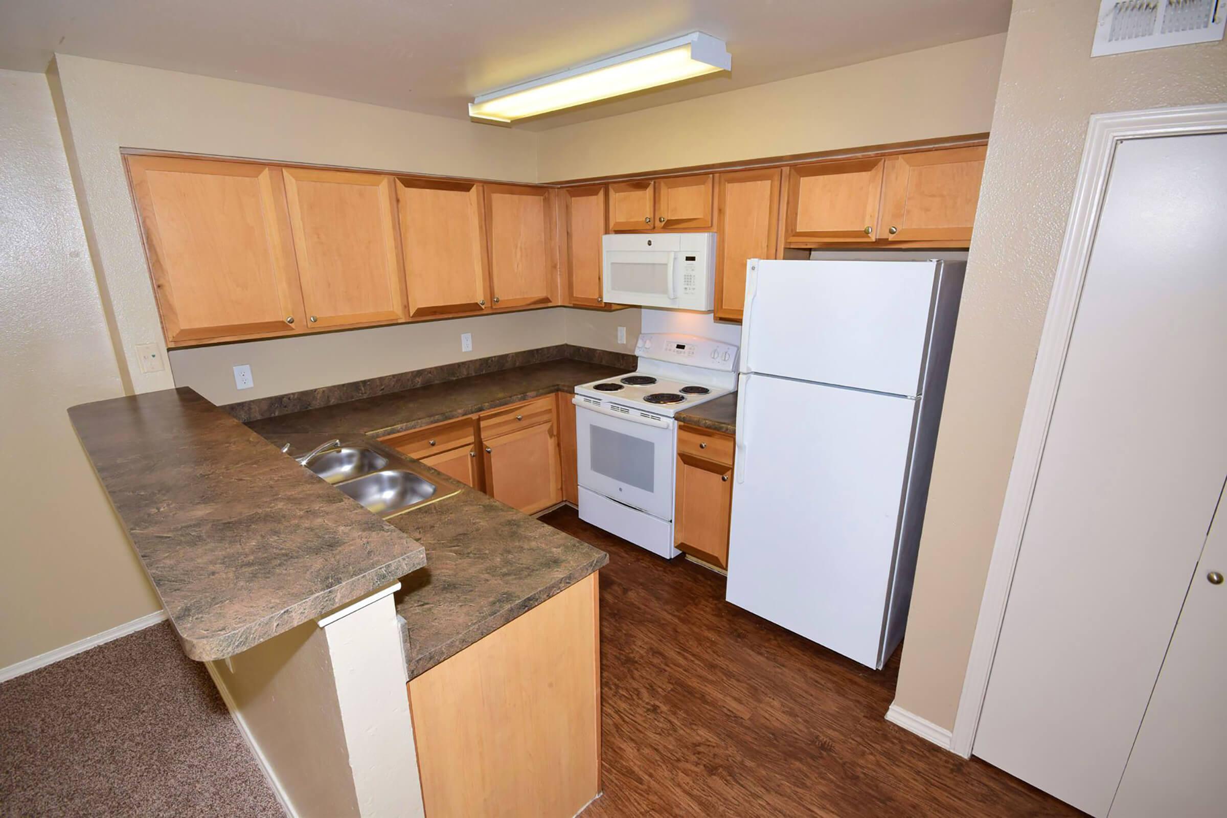 A well-lit kitchen featuring wooden cabinets, a white refrigerator, and an oven with a stovetop. The countertop is dark and includes a double sink. The walls are painted a light color, and there is a door to a pantry or closet visible. The floor has a wood-like laminate finish.