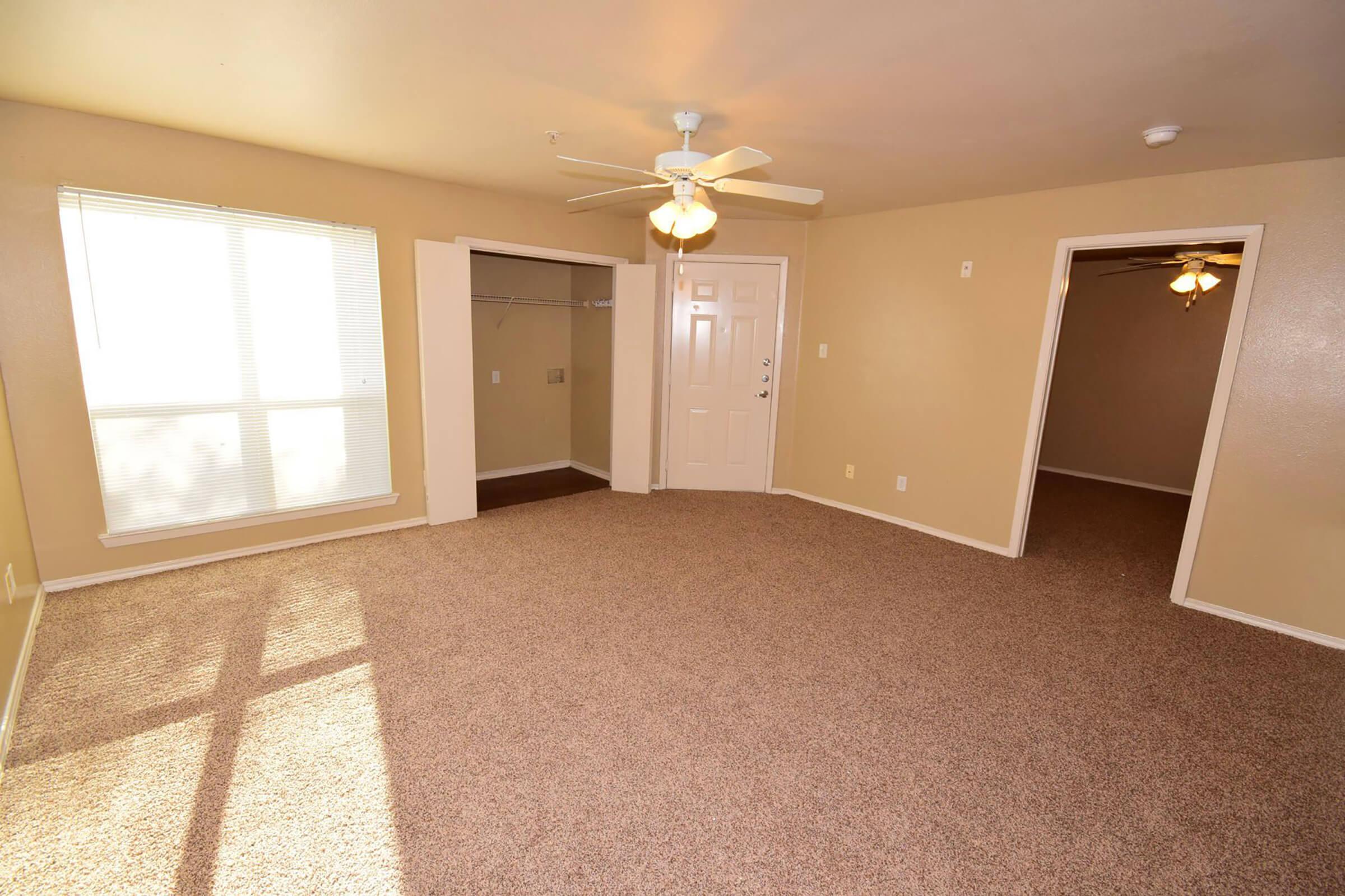 Spacious empty living room with light brown carpet, a ceiling fan, and large window letting in natural light. Features beige walls and a door leading to another room. There is a small closet area visible on the left side of the room.