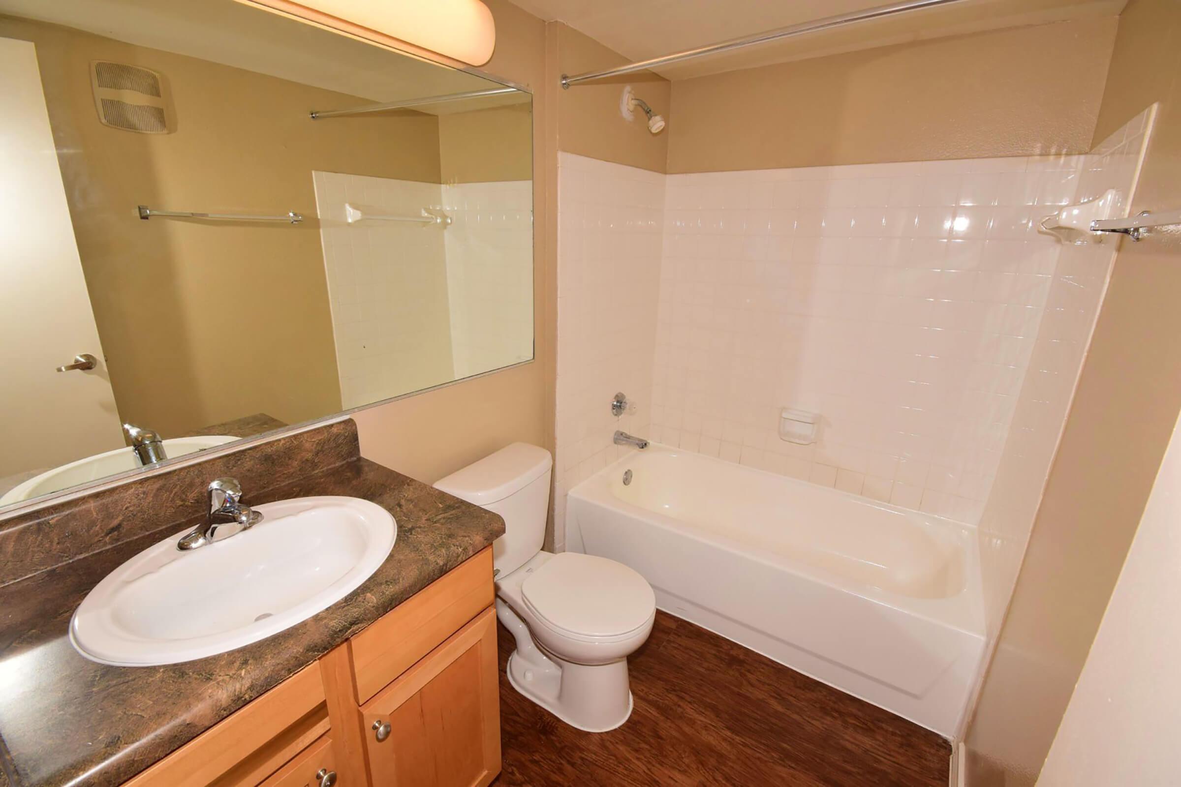 A well-lit bathroom featuring a bathtub with a shower, a sink with a mirror above it, and a toilet. The walls are painted in a light color, and the floor is a dark wood laminate. There are towel bars installed on the wall, and a wooden cabinet under the sink for storage.