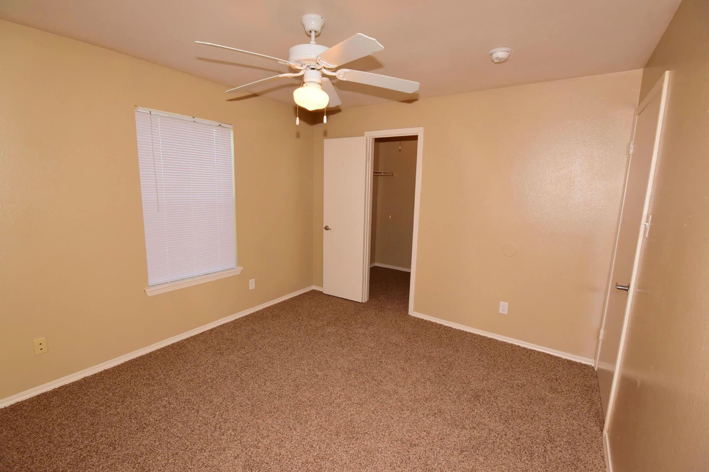 A small, empty room with beige walls and carpeted flooring. There is a single window with blinds, a ceiling fan, and a closed door leading to a closet. The space is well-lit and lacks furniture, creating an open, airy feel.