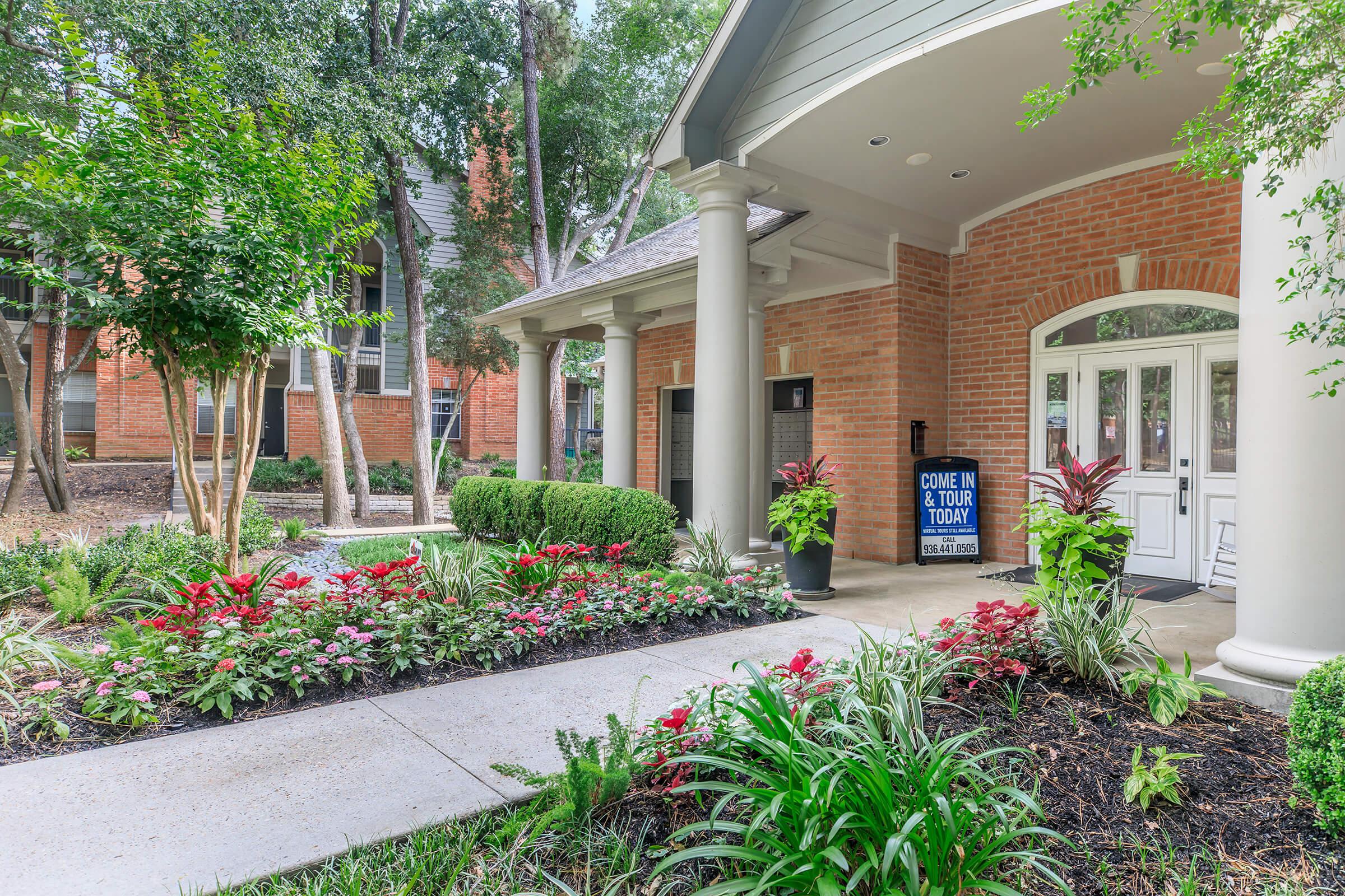 Brightly landscaped entrance to a building featuring a path lined with colorful flowers and greenery. A sign reads "Come in & tour today." The architecture includes a covered porch with white columns, surrounded by trees and well-maintained shrubs.