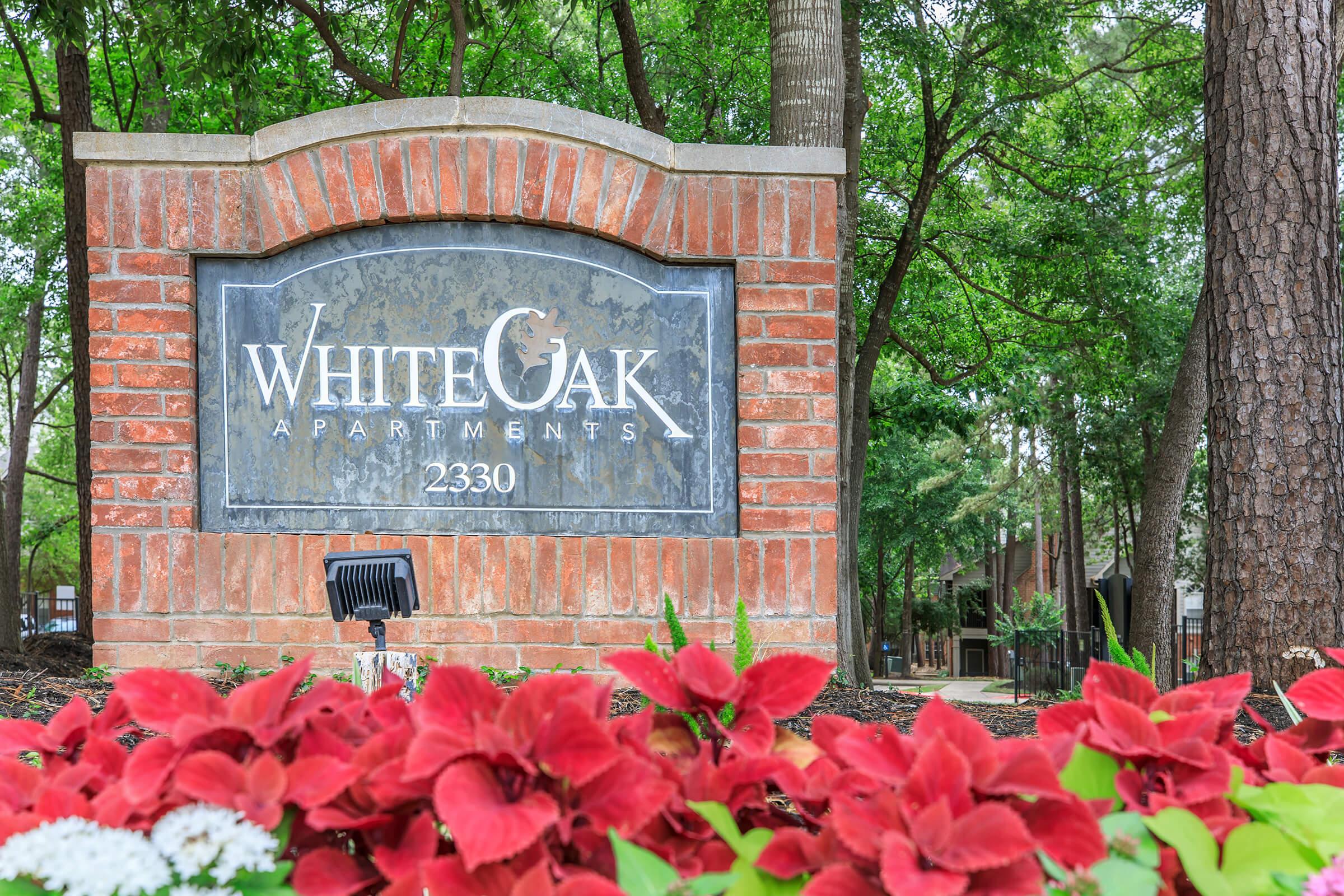 Sign for White Oak Apartments with the address 2330, surrounded by greenery and red flowers. The sign features a brick base and a decorative design, providing an inviting atmosphere for residents and visitors.
