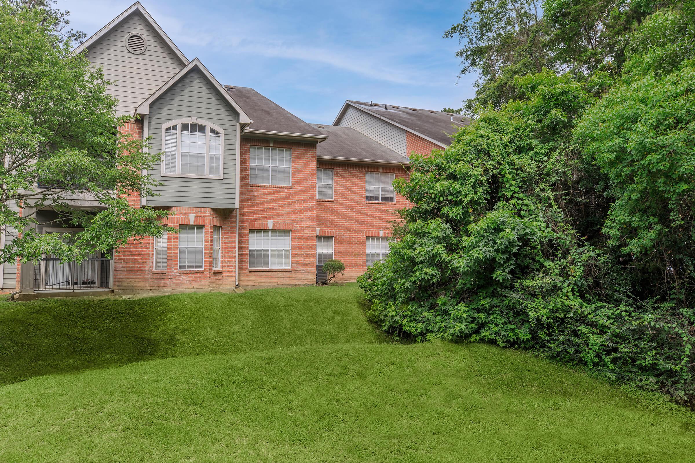 A multi-story residential building featuring a mix of red brick and gray siding, set against a backdrop of lush green foliage and a well-maintained lawn. The scene captures a peaceful, suburban environment with trees framing the structure.