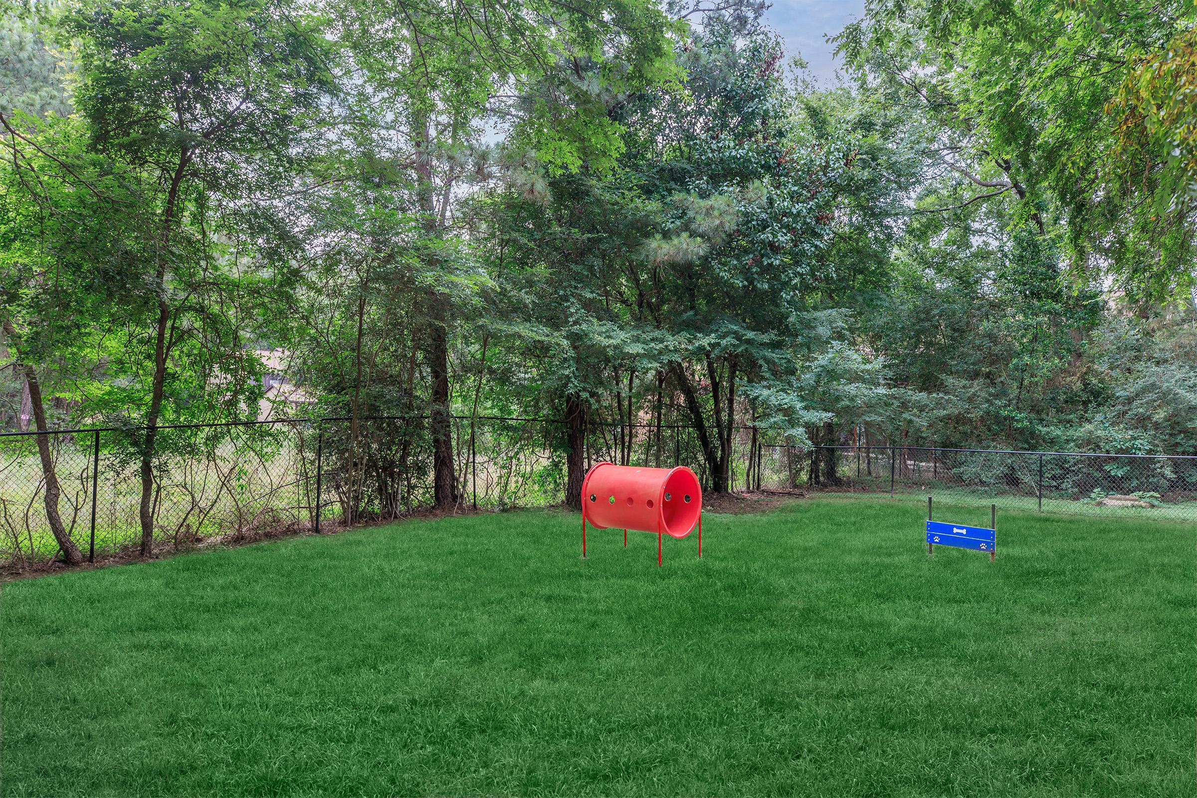 A grassy backyard surrounded by trees, featuring a red tunnel-like structure for pets and a blue sign, all enclosed by a fence. The scene is tranquil, with natural greenery providing a peaceful atmosphere.