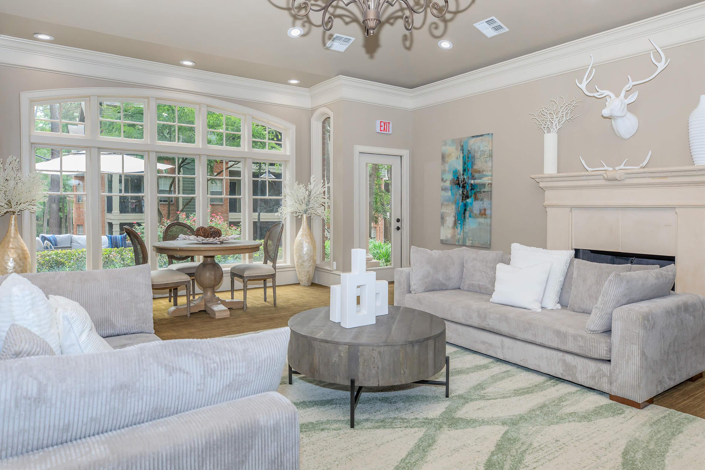 A bright and modern living room featuring a plush gray sofa and a round coffee table. The walls are light-colored, adorned with decorative elements. A large window offers a view of the outdoors, and there are decorative antlers and vases on the mantle above the fireplace. A dining area is visible in the background.