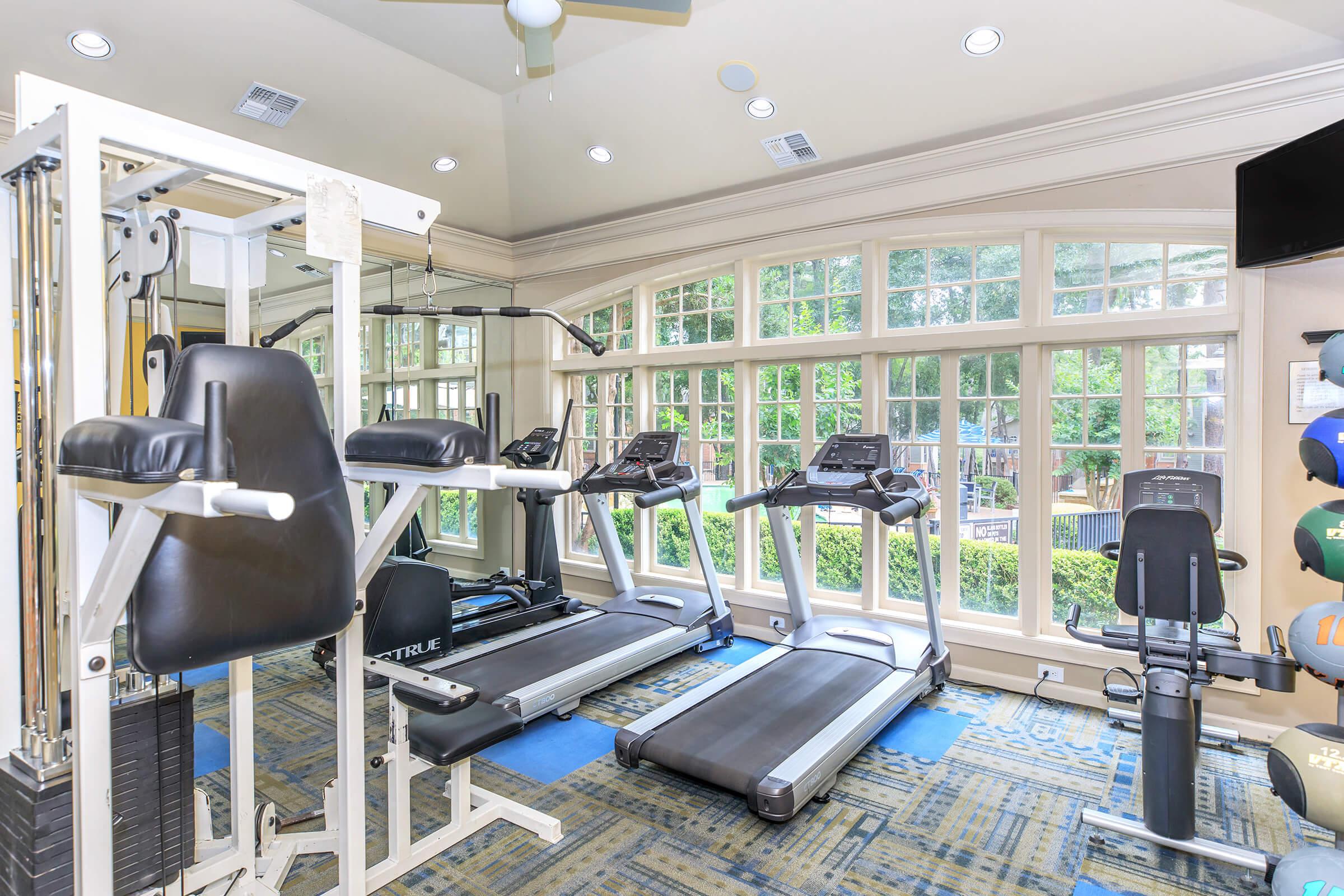 A spacious fitness room with large windows letting in natural light. It features various gym equipment, including treadmills, strength training machines, and a stationary bike. The floor is carpeted, and there is a green view from the windows, creating an inviting workout environment.