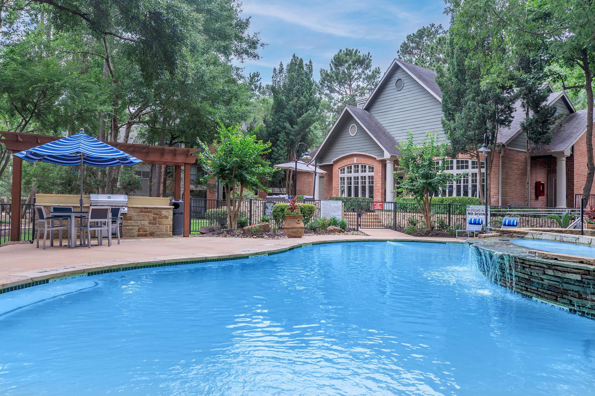 A serene outdoor swimming pool with a clear blue surface, surrounded by lush greenery and a patio area featuring a striped umbrella and seating. In the background, a cozy brick building with large windows adds to the inviting atmosphere, creating a perfect setting for relaxation.