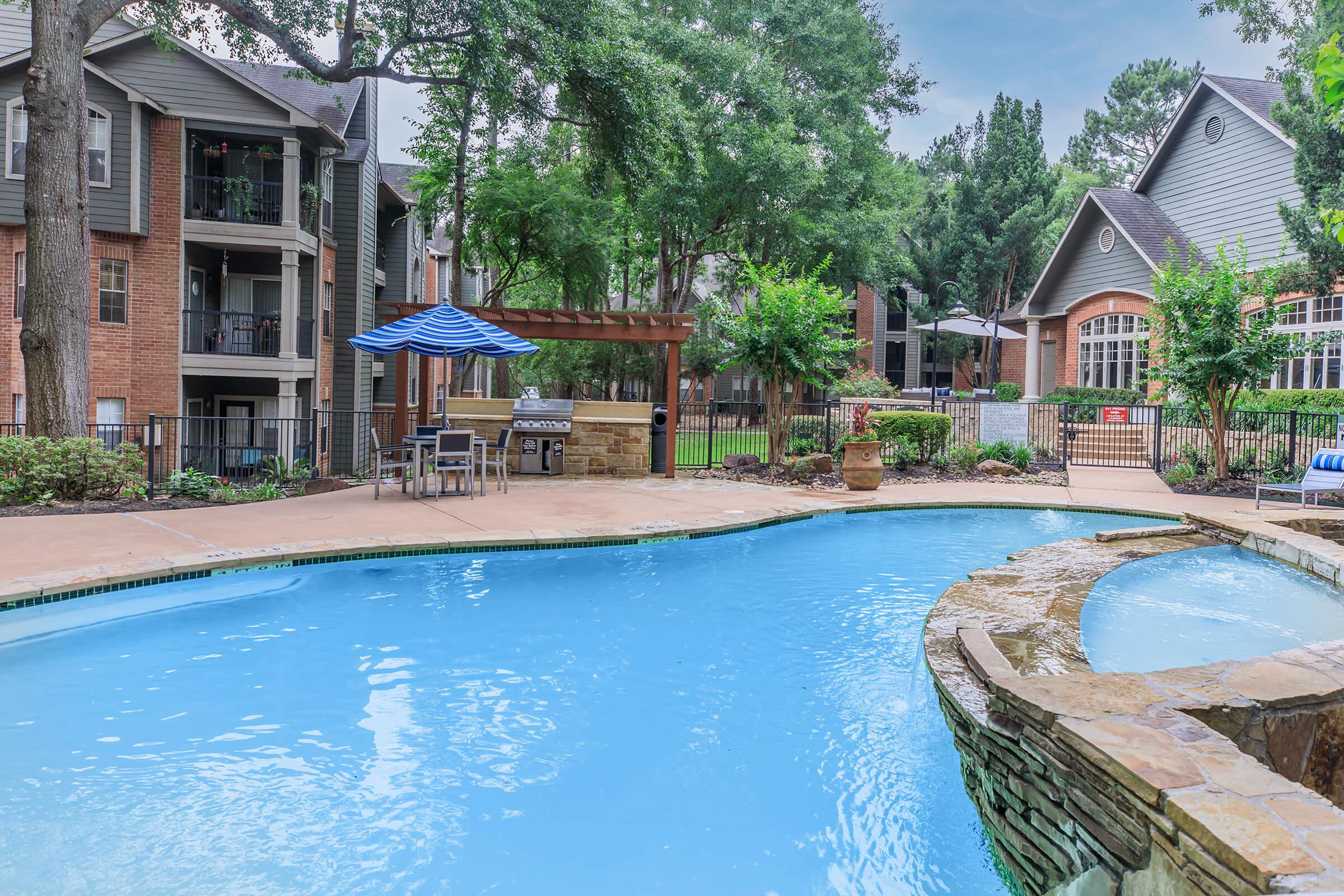 A serene swimming pool surrounded by lush greenery and trees, with a shaded seating area featuring a blue and white striped umbrella. Nearby, there are apartment buildings with brick exteriors and an inviting entrance. The atmosphere is relaxed and perfect for leisure activities.