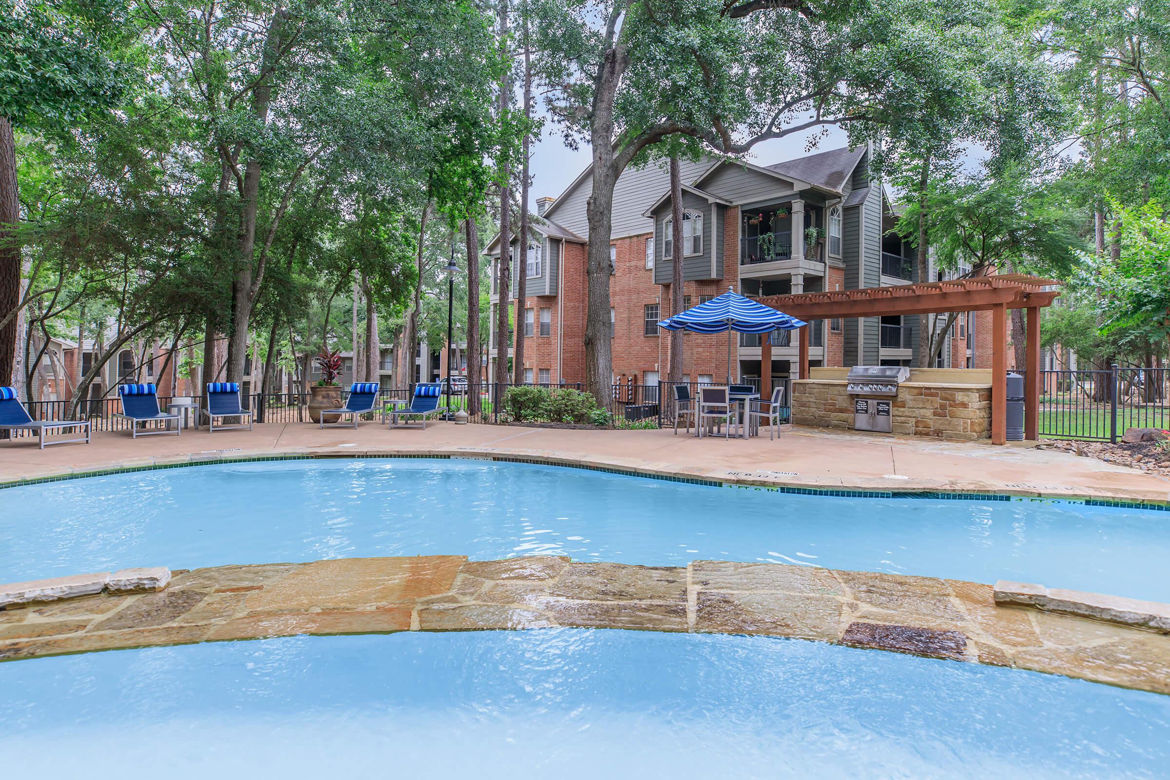 A serene outdoor pool area surrounded by lush trees, featuring a curved pool with a stone edge. In the background, there are multi-story apartment buildings with balconies. Several lounge chairs are arranged by the poolside, and a shaded patio area with tables and chairs is visible under a blue umbrella.