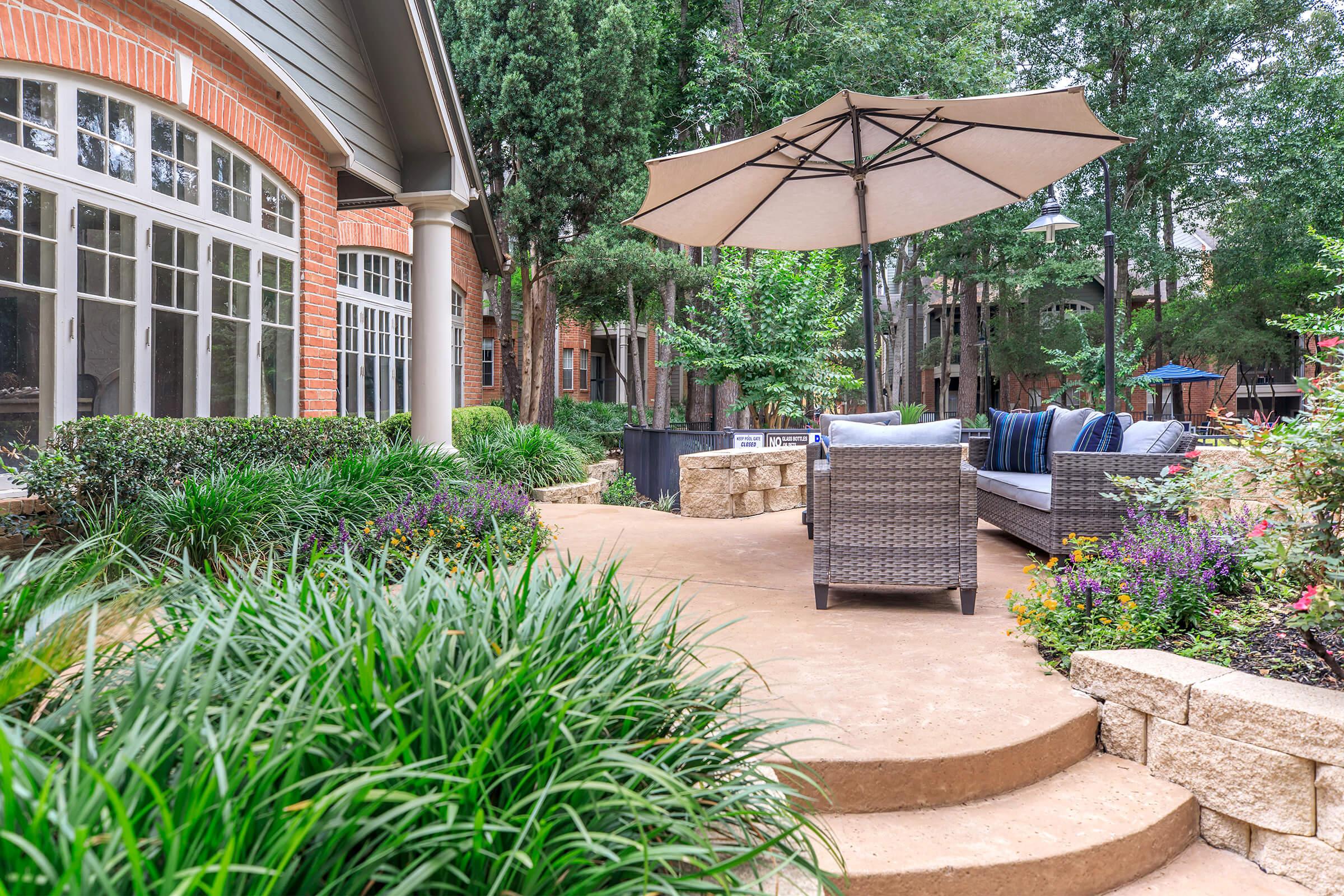 A cozy outdoor space featuring a patio with a comfortable seating area, including a sectional sofa and an umbrella for shade. Lush greenery surrounds the path, with colorful flowers and well-maintained landscaping, creating a serene and inviting atmosphere.