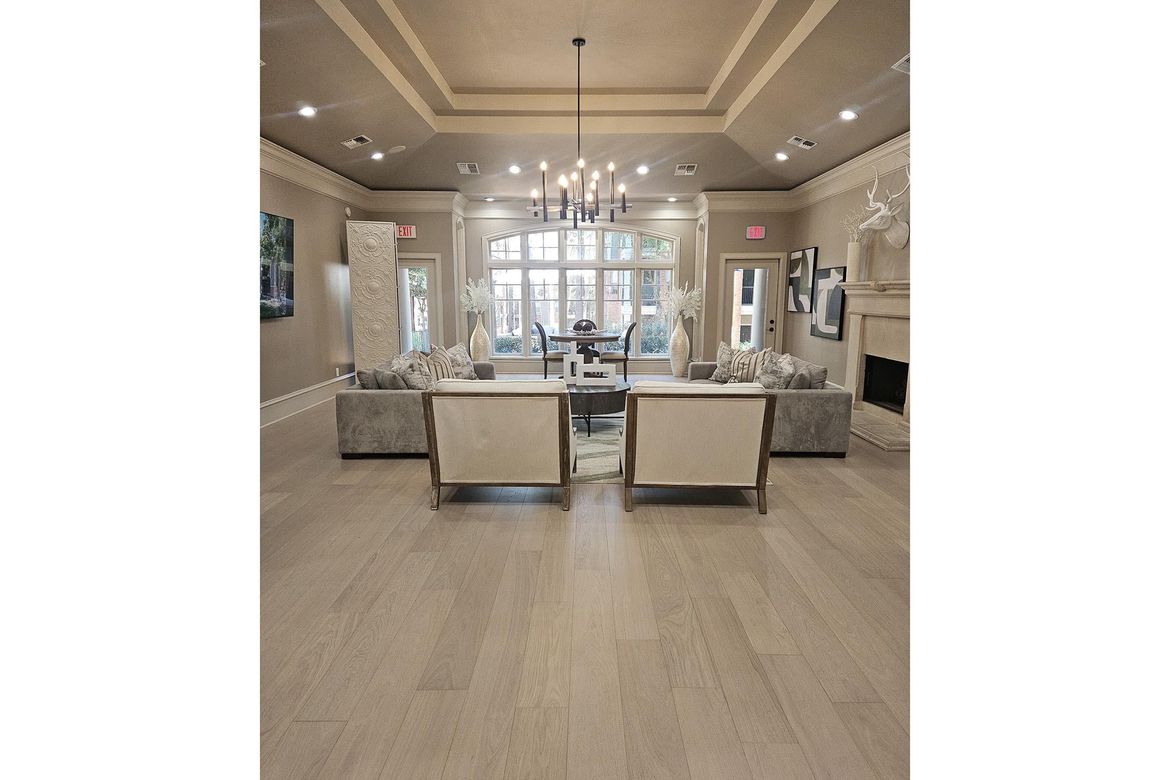 Spacious living room with light hardwood floors, featuring two cream-colored armchairs facing a soft gray couch. The room is well-lit with a chandelier and large windows, showcasing a modern decor style, including artwork and decorative accents. A dining area is visible in the background.