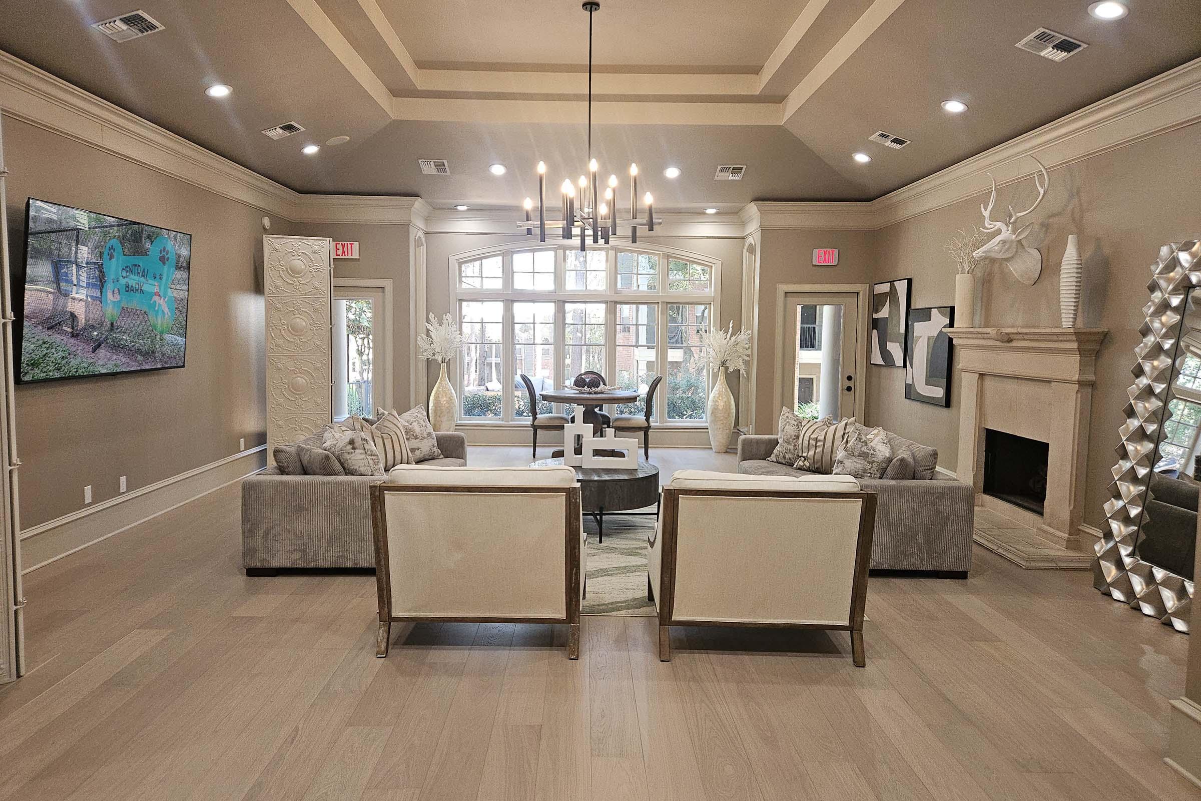 A spacious living room featuring a stylish seating arrangement with beige and gray sofas, a coffee table, and a modern chandelier. Large windows allow natural light in, while decorative elements like wall art, a fireplace, and a unique mirror enhance the elegant decor. The overall ambiance is welcoming and contemporary.