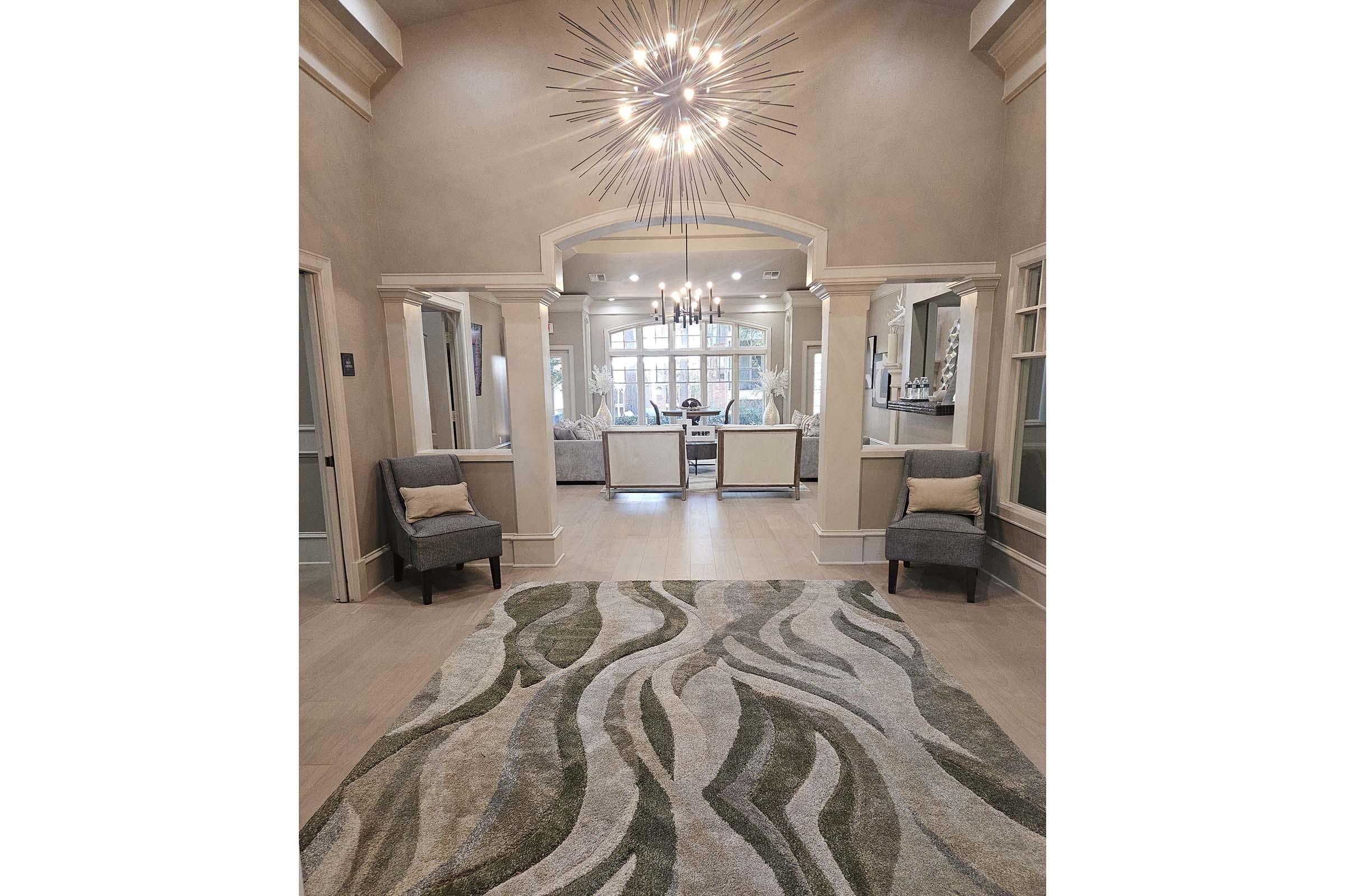 A spacious entryway featuring a large decorative area rug with wavy patterns in neutral tones. Two stylish armchairs are positioned on either side, with a modern chandelier hanging overhead. The background reveals a well-lit living area with elegant furniture and a warm atmosphere.