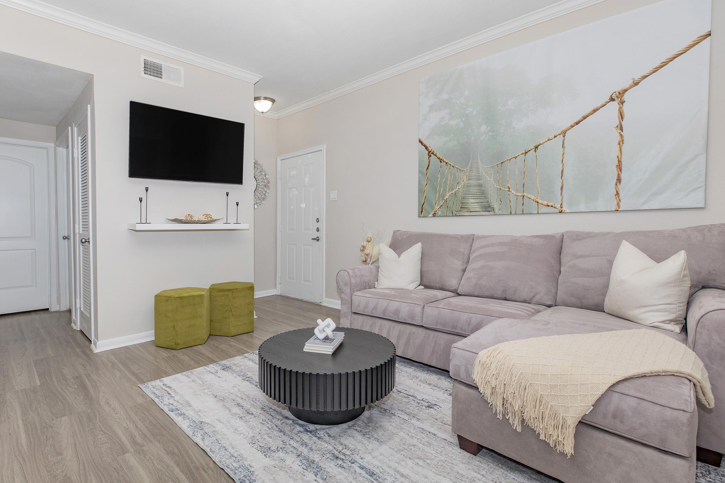 Cozy living room featuring a light beige sectional sofa with white pillows, a round black coffee table, and a decorative rug. A large wall art piece of a rope bridge is above, and a TV mounted on the wall adds functionality. Two green ottomans and soft lighting create a warm, inviting atmosphere.