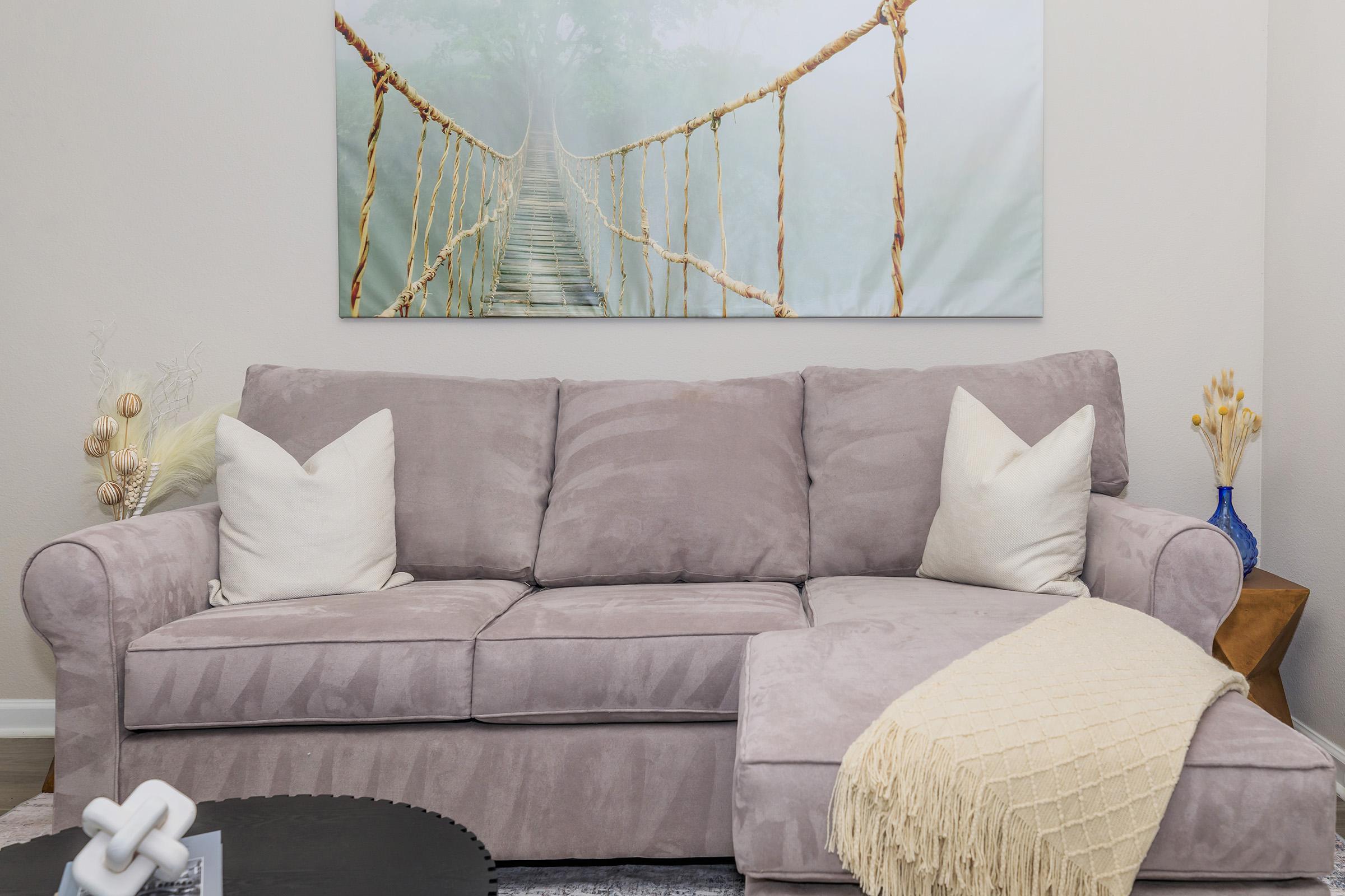 A cozy living room featuring a light gray sectional couch with two white pillows. A soft cream throw blanket drapes over one side of the sofa. In the background, there is a large artwork depicting a foggy forest with a rope bridge, enhancing the serene atmosphere of the space.