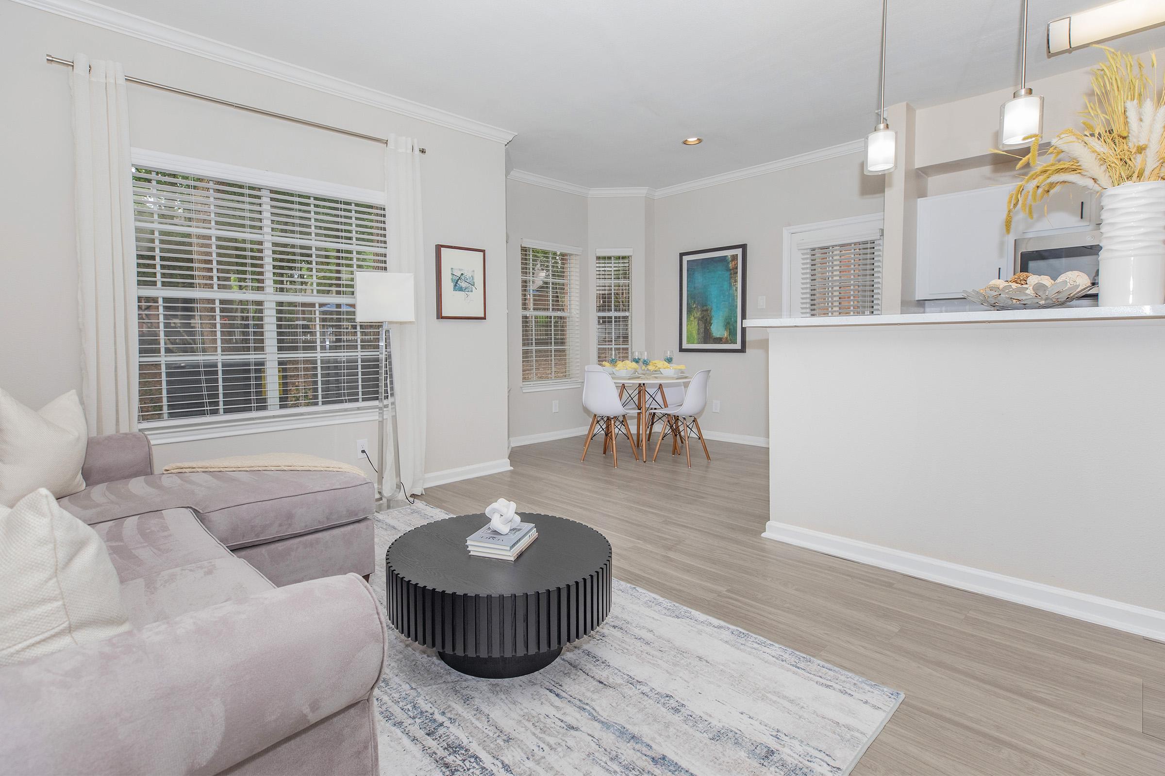 A cozy living space featuring a light gray couch, a round black coffee table, and a dining area with a white table and chairs. Natural light streams through the windows, complemented by light curtains and decorative artwork on the walls. The flooring is light wood, creating a warm and inviting atmosphere.