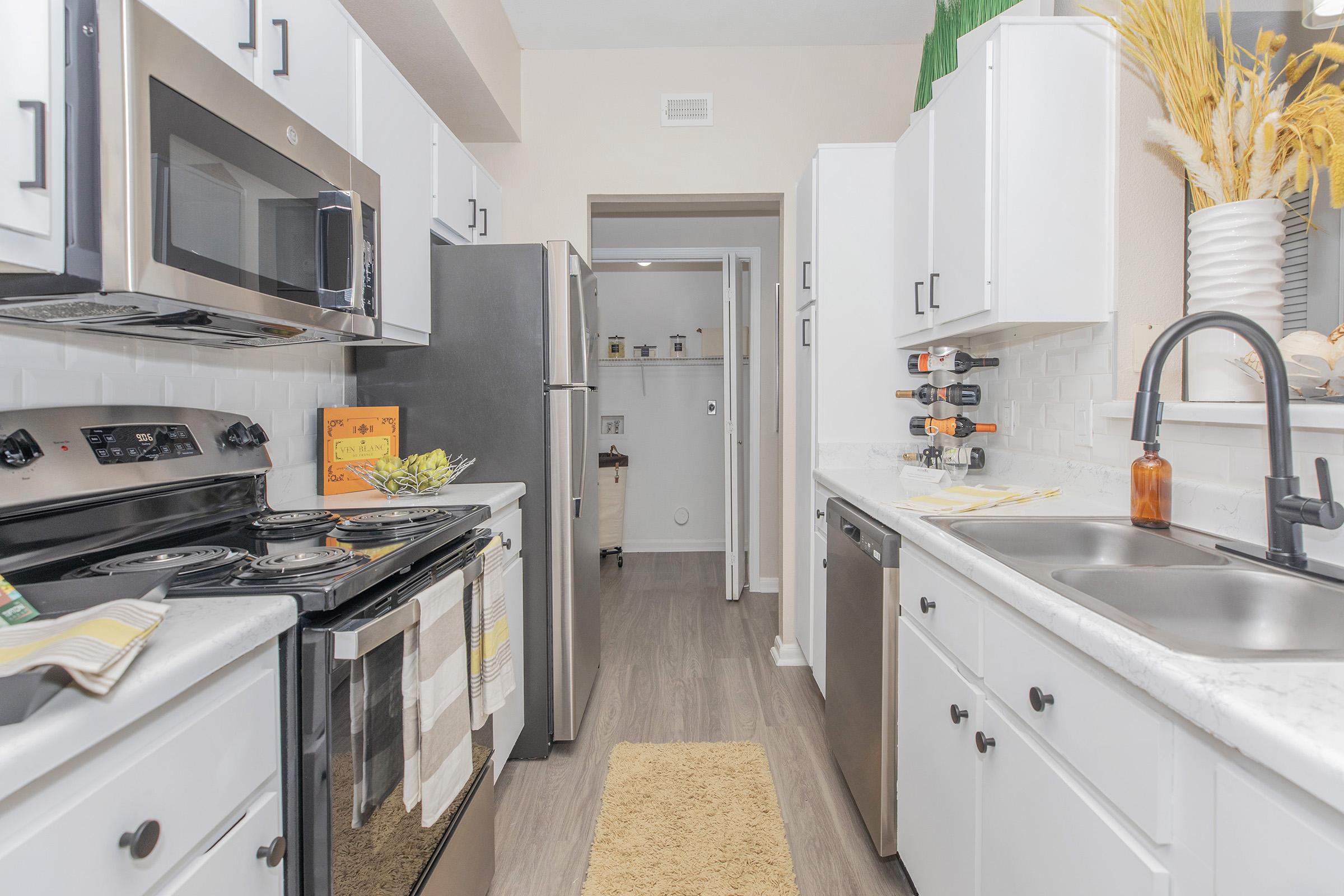 A modern kitchen featuring white cabinets, stainless steel appliances, and a marble countertop. There’s a stove, microwave, and refrigerator, alongside a sink with a stylish faucet. The kitchen has decorative items, including a vase with dried plants, and a rug on the floor, creating a warm and inviting atmosphere.
