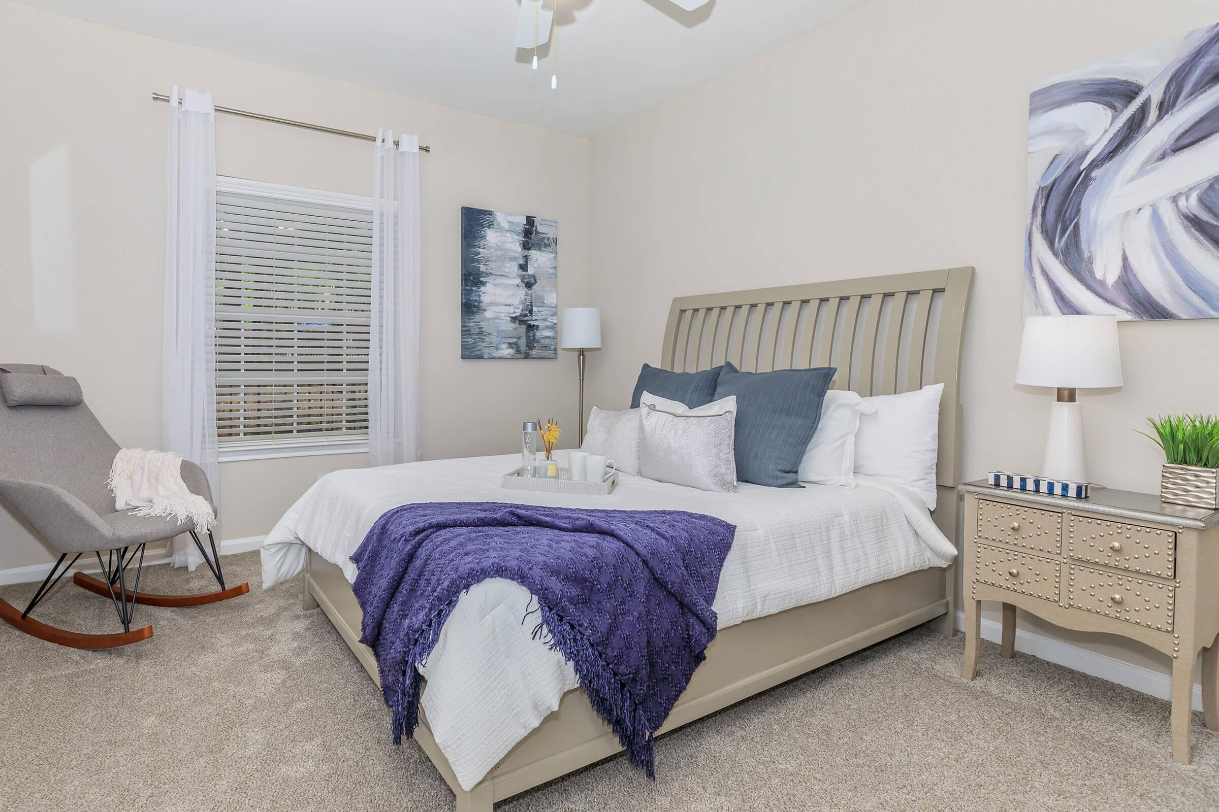 A cozy bedroom featuring a large bed with white linens and a purple throw blanket. There's a rocking chair in the corner, a bedside table with decorative items, and two lamps for lighting. The walls are painted a light color, and a piece of abstract art hangs above the bed. Natural light filters through a window with sheer curtains.