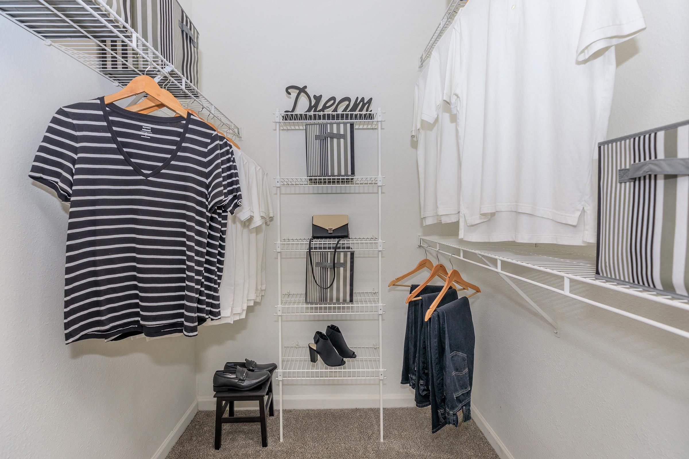 A neatly organized closet featuring striped and solid white t-shirts hanging on wire shelves. A small chair is placed next to a set of black shoes and a rack displaying denim pants. A decorative sign reading "Dream" is positioned on the shelf, alongside black and white striped storage boxes.