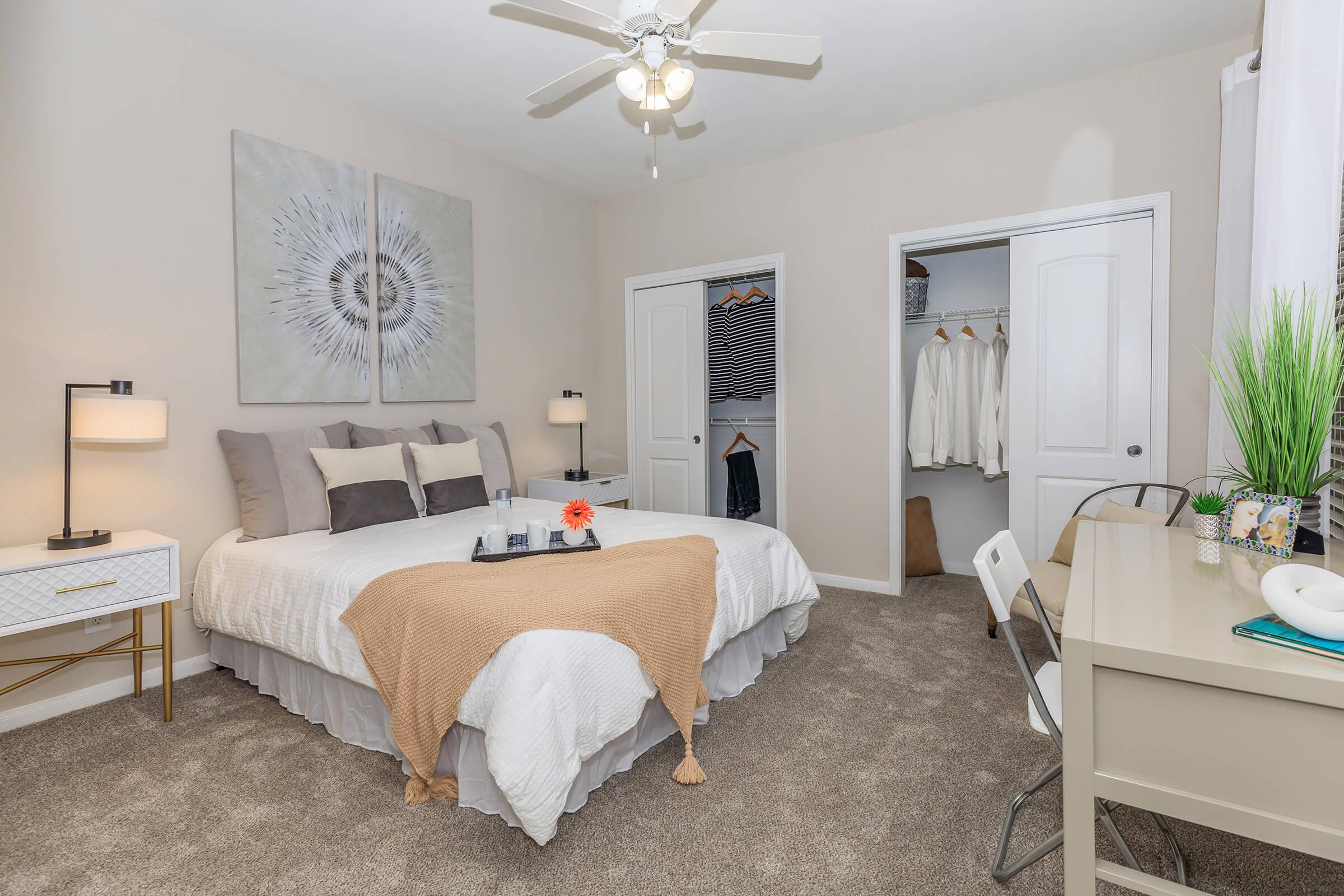 A cozy bedroom featuring a comfortable bed with decorative pillows and a light blanket. A nightstand with a lamp sits beside the bed. A desk and chair are positioned near a window, with a closet visible, containing clothes. Light-colored walls and carpet create a warm atmosphere.