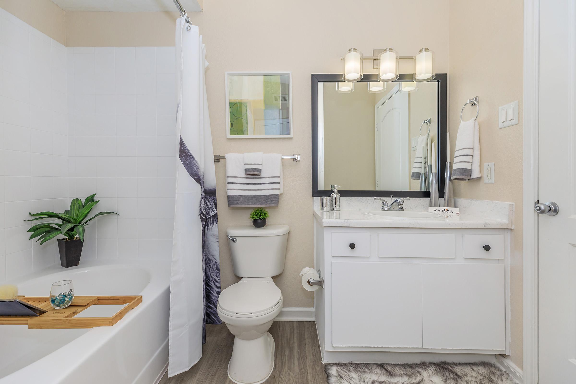A modern bathroom featuring a white bathtub with a wooden tray, a white toilet, and a sink with a large mirror above it. Stylish light fixtures hang over the mirror. The walls are painted a light beige, and a small potted plant adds a touch of greenery to the space. Towels are neatly arranged on a rack.