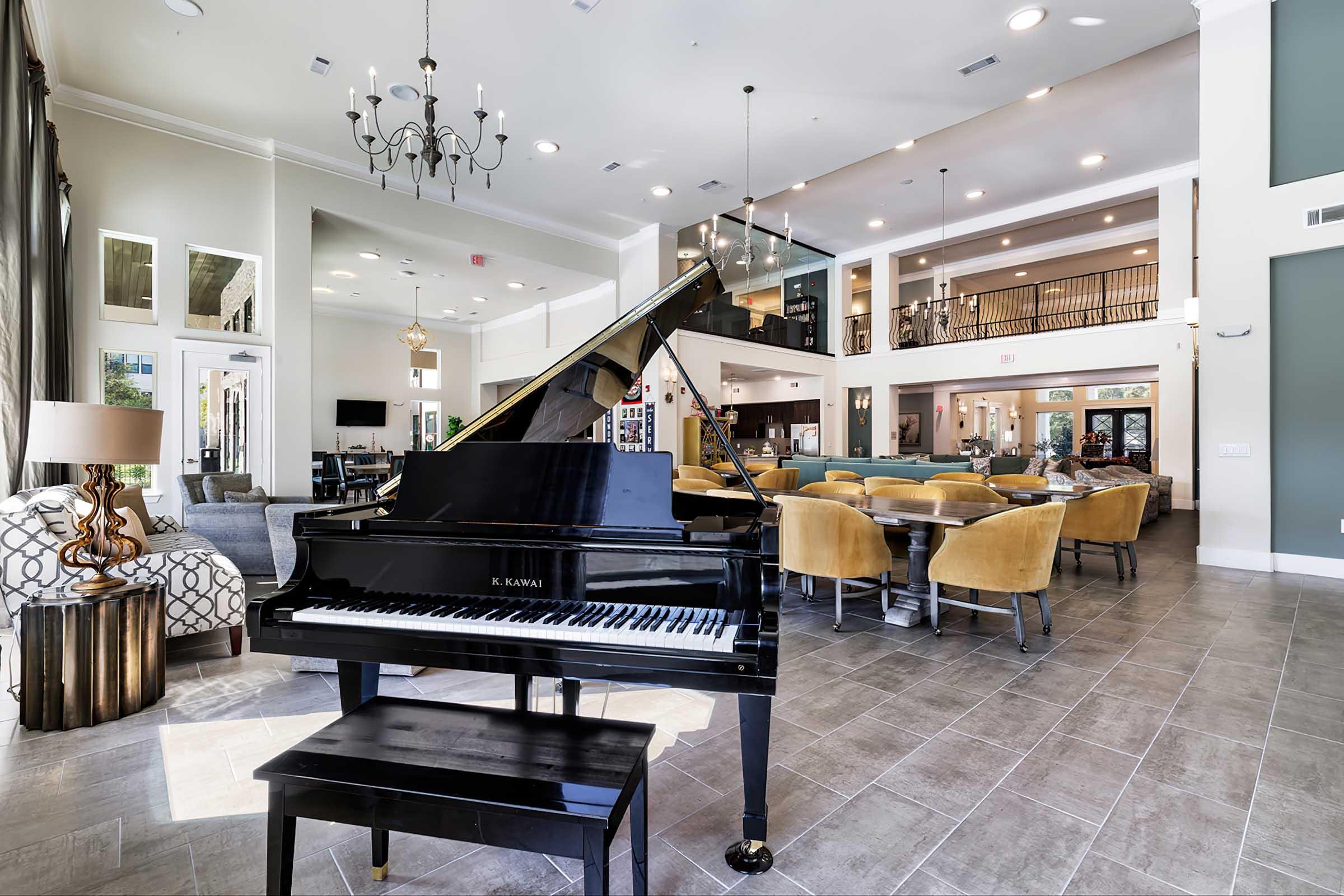 A spacious and elegant common area featuring a grand piano in the foreground. The room has high ceilings, large windows, and modern furnishings, including comfortable seating and a dining area. Decorative chandeliers hang from the ceiling, and the walls are adorned with art.