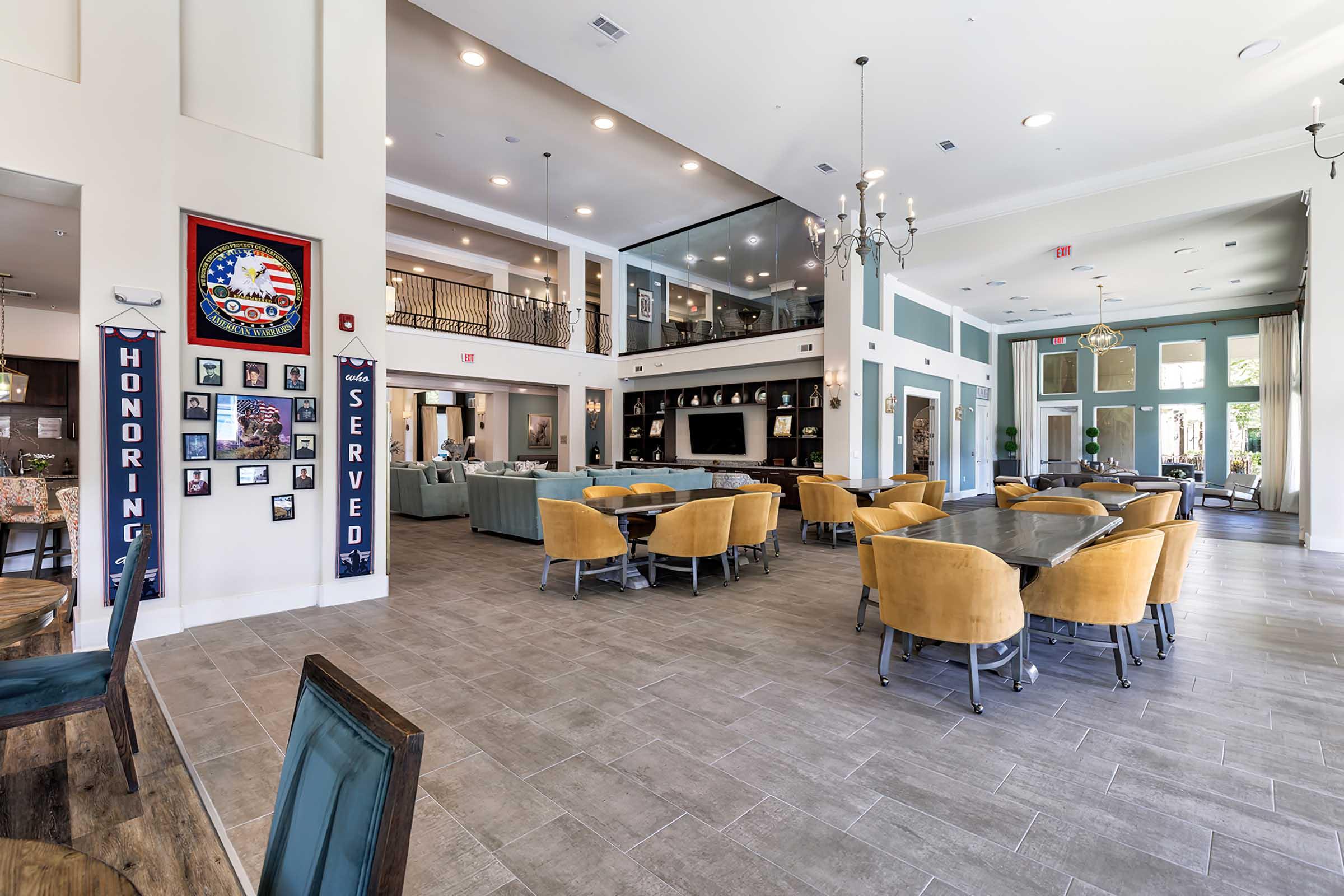 A spacious and modern common area featuring dining tables with yellow chairs, comfortable seating arrangements, and large windows. The walls display various framed photographs and an American flag-themed decoration. Nearby, there are chandeliers and a cozy atmosphere.