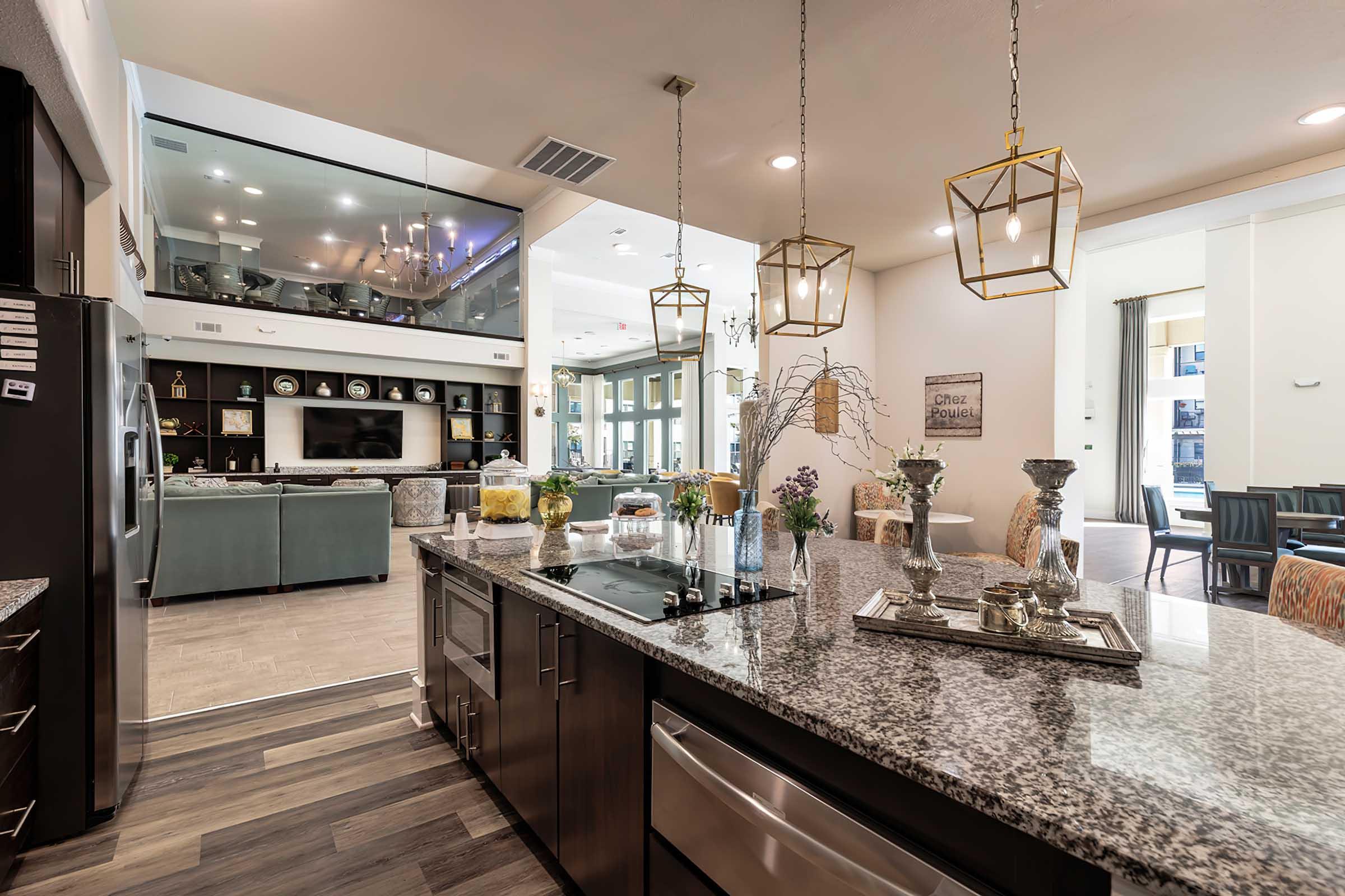 A modern kitchen featuring dark cabinets, a granite countertop, and pendant lighting. There are decorative vases and candles on the countertop, with a view of a stylish dining area and living room in the background. The space is bright and airy, decorated with contemporary furnishings.