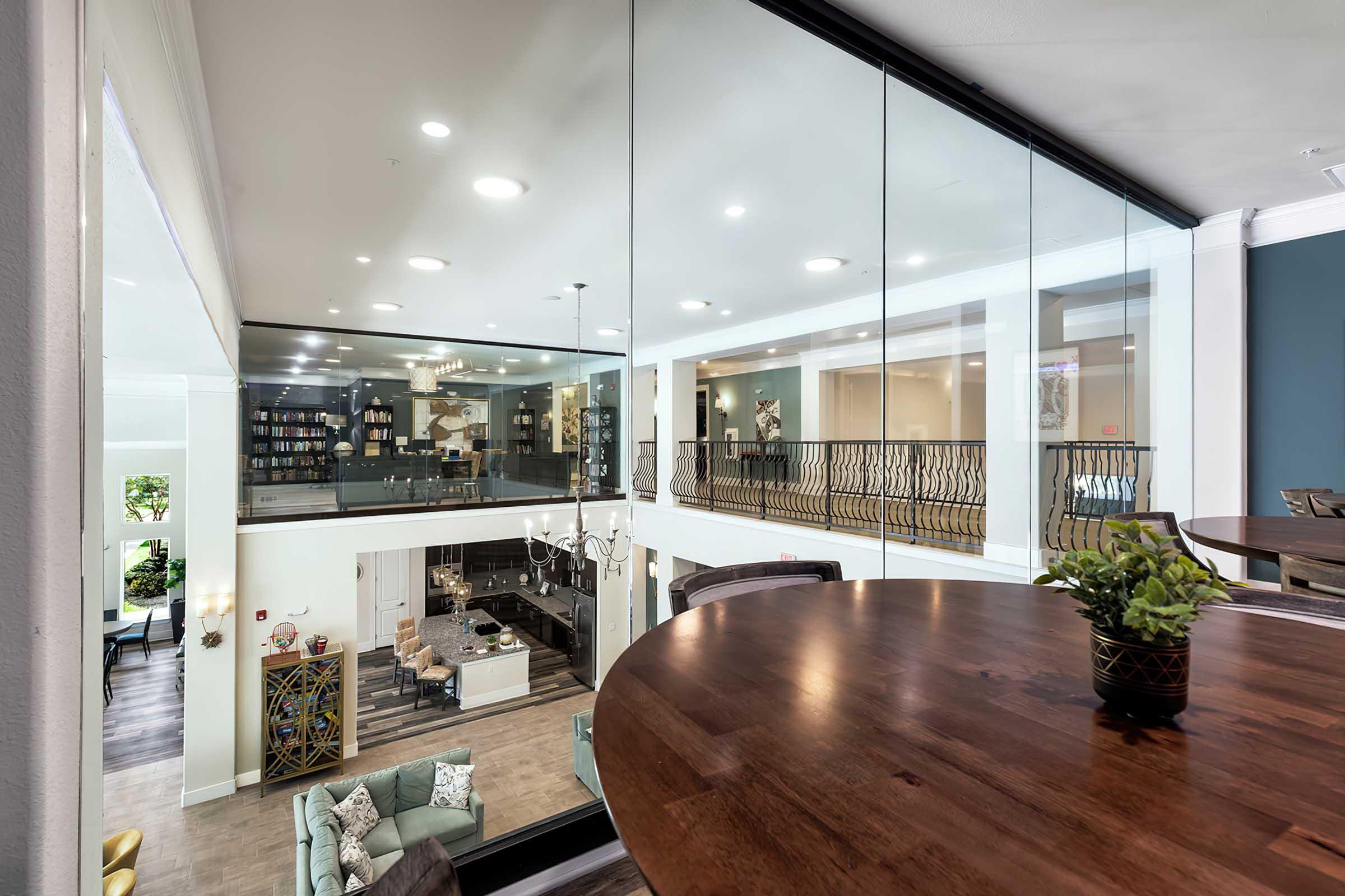 Interior view of a modern, spacious common area showcasing a second-level balcony with glass railings, overlooking a stylishly furnished lounge and dining space below. Natural light streams in through large windows, highlighting contemporary decor and a cozy atmosphere.