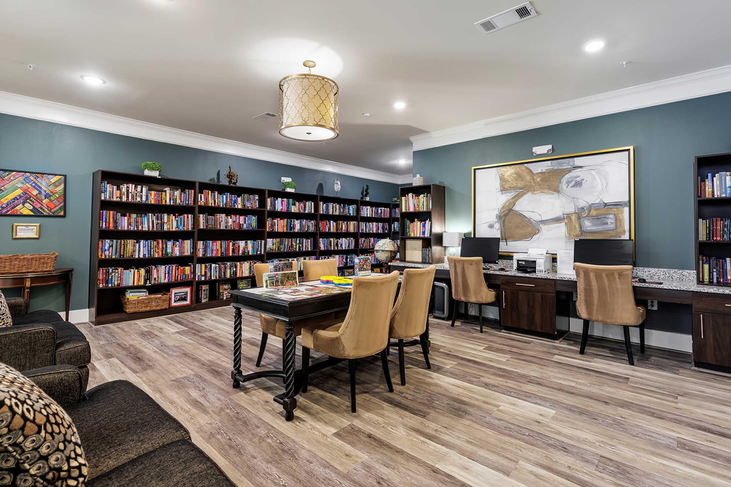 A cozy library room featuring dark blue-green walls, large bookshelves filled with colorful books, a comfortable seating area with a sofa, and a desk with chairs for reading or working. The room is well-lit with overhead lights and decorated with artwork.