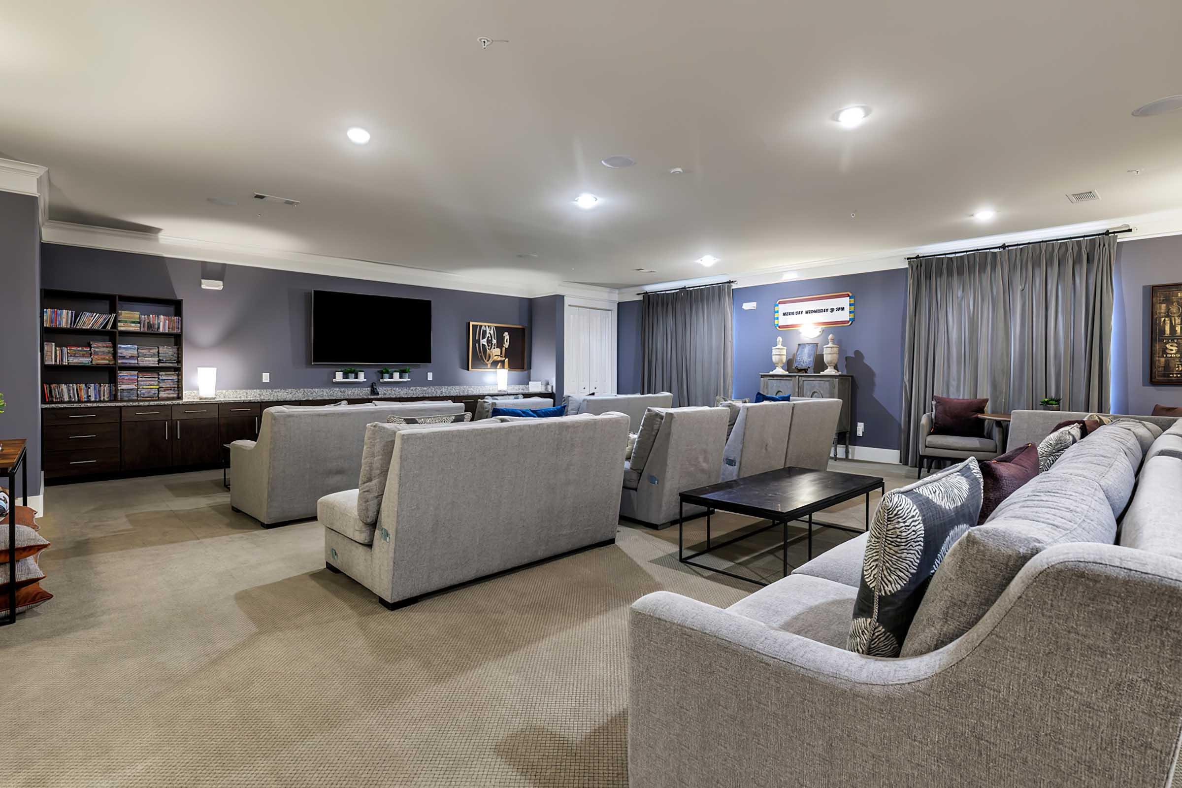 A spacious, modern living room featuring several light gray sofas arranged around a central coffee table, with a large flat-screen TV mounted on the wall. The room is illuminated with soft lighting, and decorative elements include artwork on the walls and vases on a side table. Large windows are covered with curtains, contributing to a cozy ambiance.