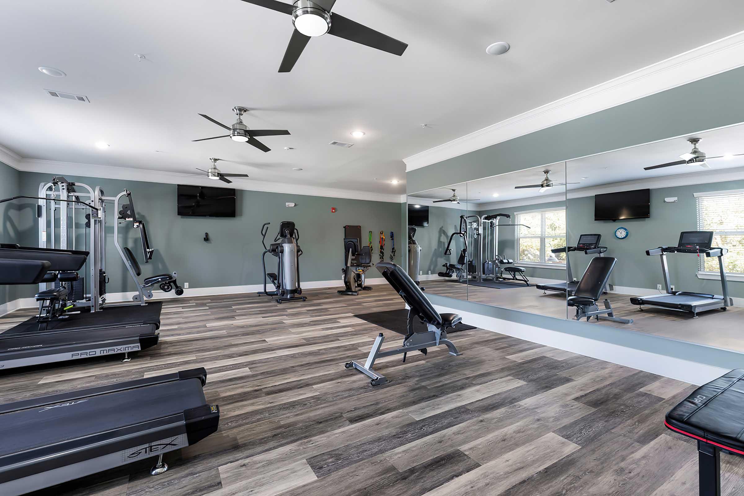 A modern fitness center featuring exercise equipment such as treadmills, weight machines, and benches. The room has wooden flooring, mirrors on one wall, and several ceiling fans. Large windows provide natural light.