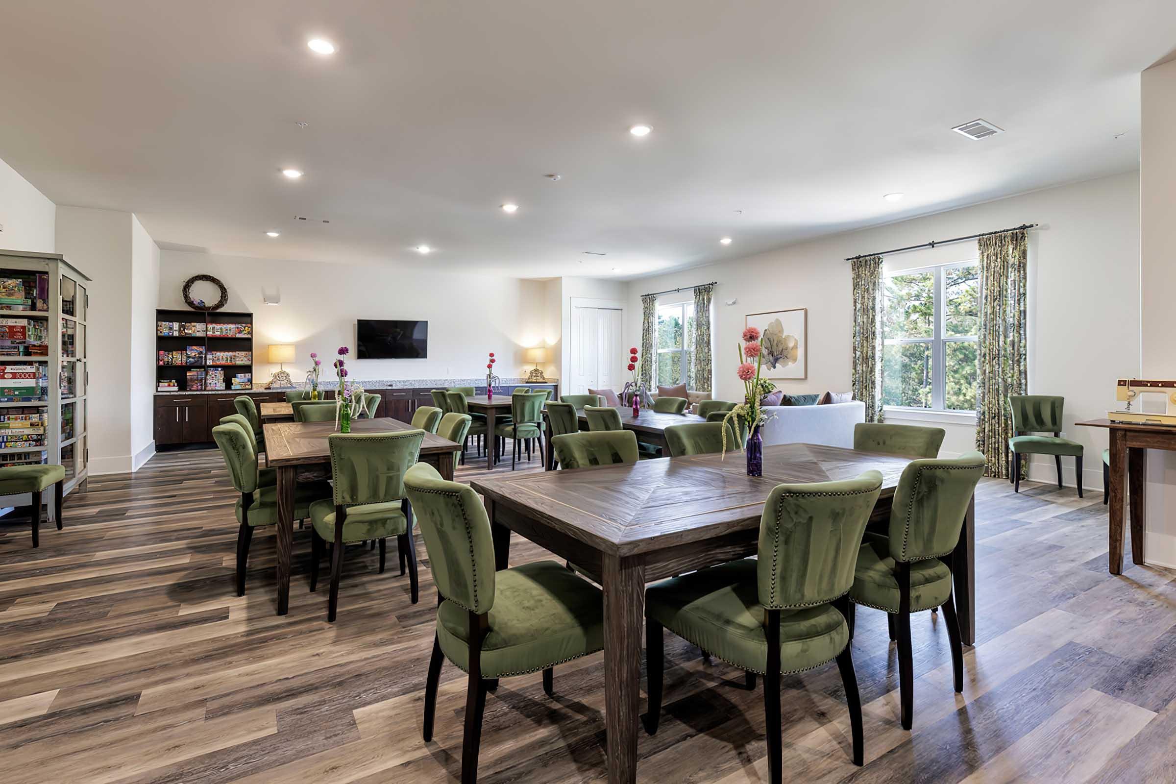 Interior of a spacious, well-lit lounge area featuring wooden tables and green upholstered chairs. There are potted plants and floral arrangements on the tables, a television mounted on the wall, and shelves filled with games. Large windows allow natural light to fill the room, enhancing the inviting atmosphere.