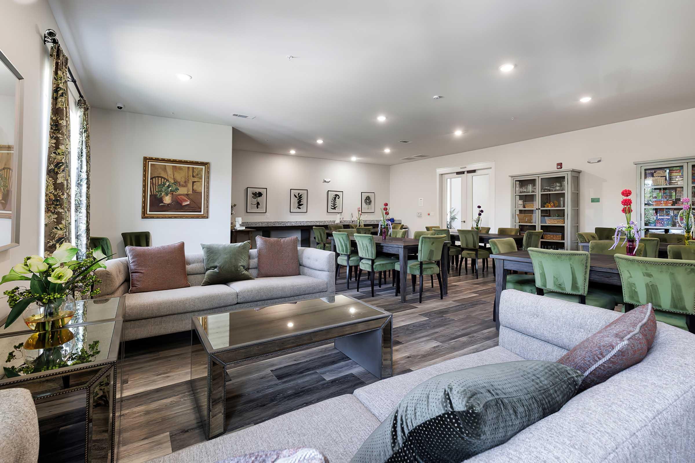 A spacious interior of a common area featuring a seating arrangement with gray sofas and a glass coffee table. The room is well-lit with large windows, and there are green dining chairs around tables in the background. Decorative plants and art pieces adorn the walls, creating a welcoming atmosphere.