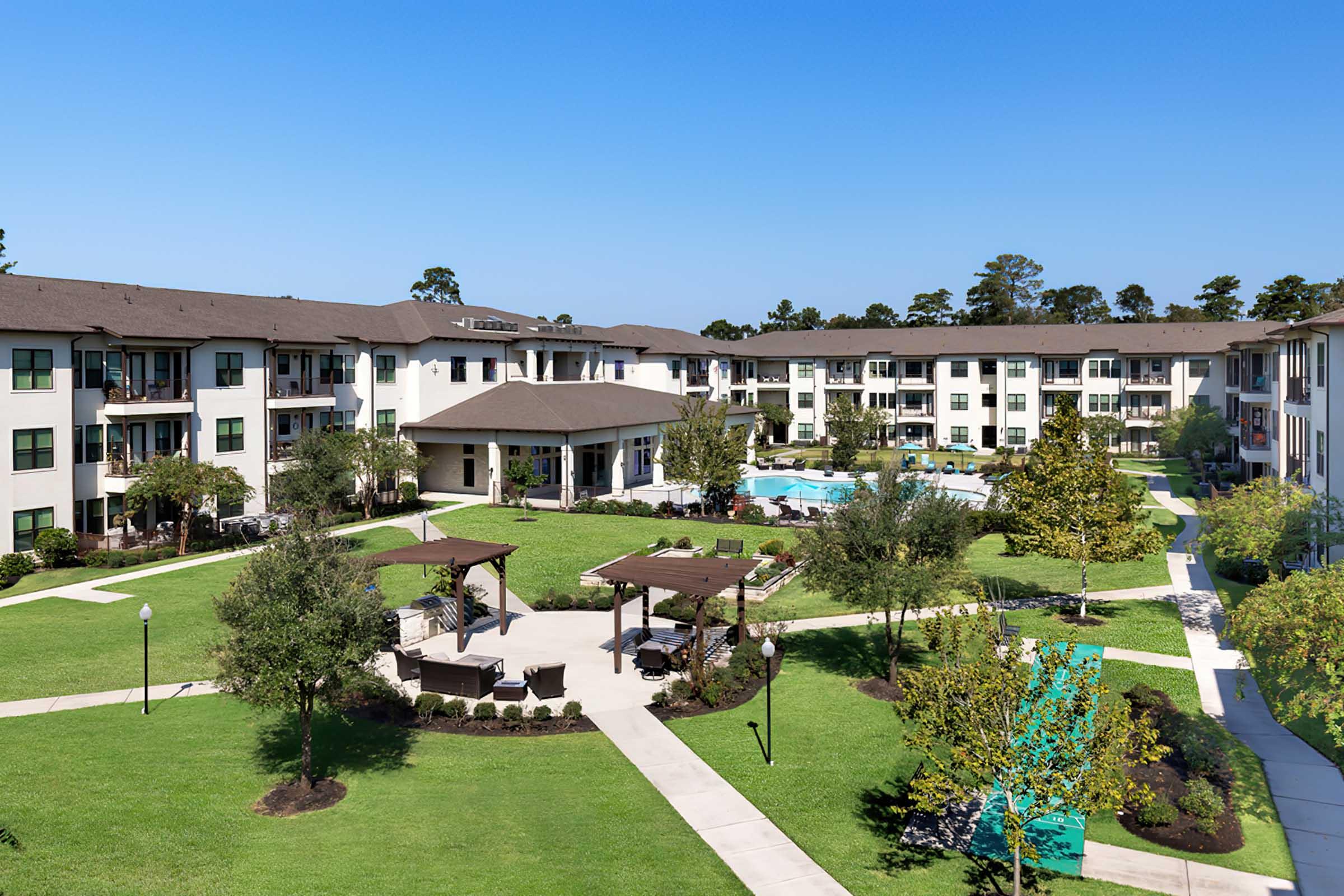 Aerial view of a modern apartment complex featuring multiple buildings surrounding a landscaped courtyard with trees, lounge areas, and a swimming pool. The sky is clear and blue.